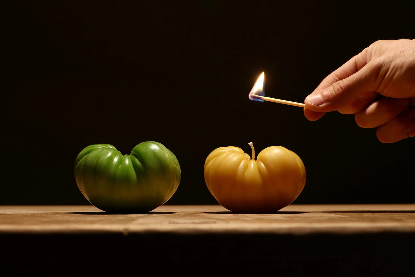 A green tomato next to a yellow tomato beeswax candle on a wooden surface with a hand holding a match.
