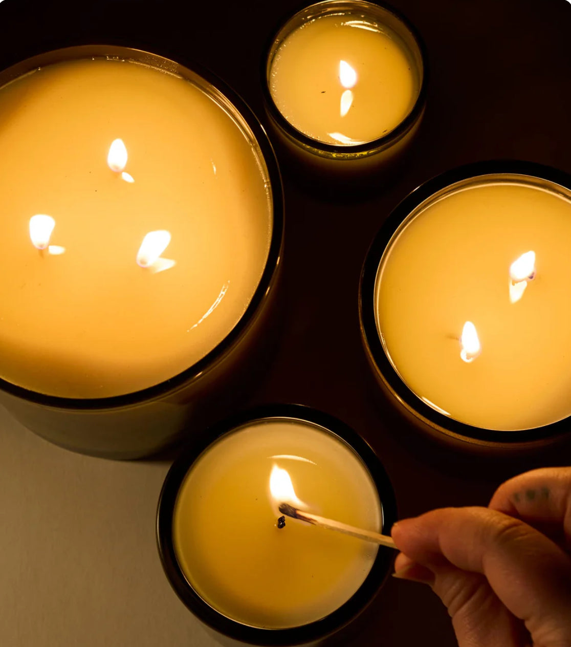 Candle holders with lit candles on a dark surface, one being lit with a match.