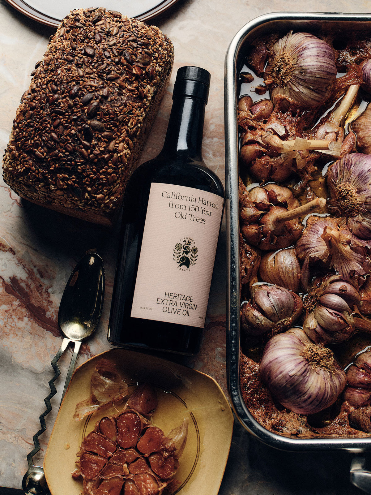 Bottle of extra virgin olive oil made from hand-harvested heirloom olives, with roasted garlic and bread on a marble surface