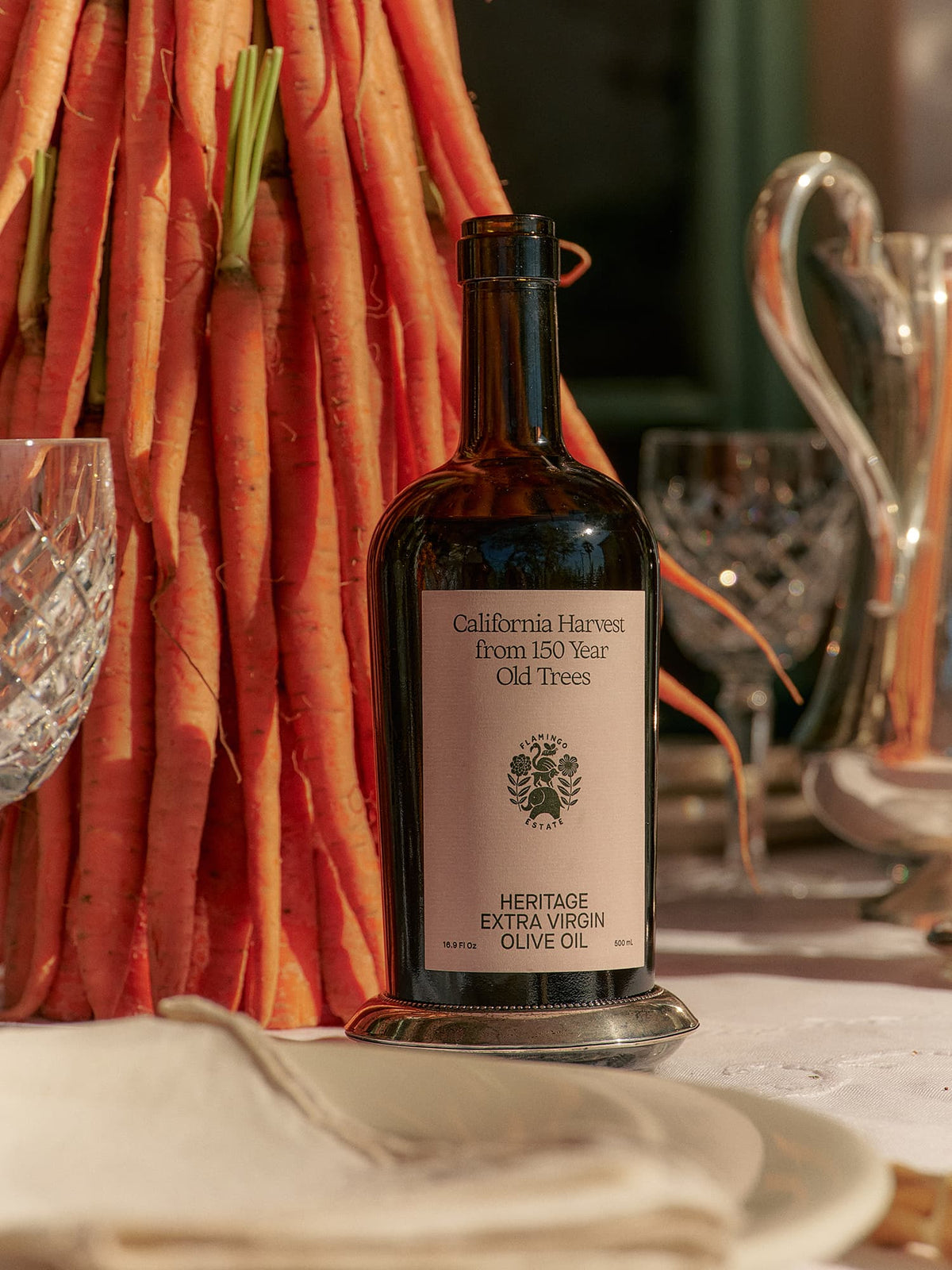 A bottle of olive oil with a label that reads 'California Harvest from 150 Year Old Trees', 'Heritage Extra Virgin Olive oil' from Flamingo Estate, on a table with decorative elements