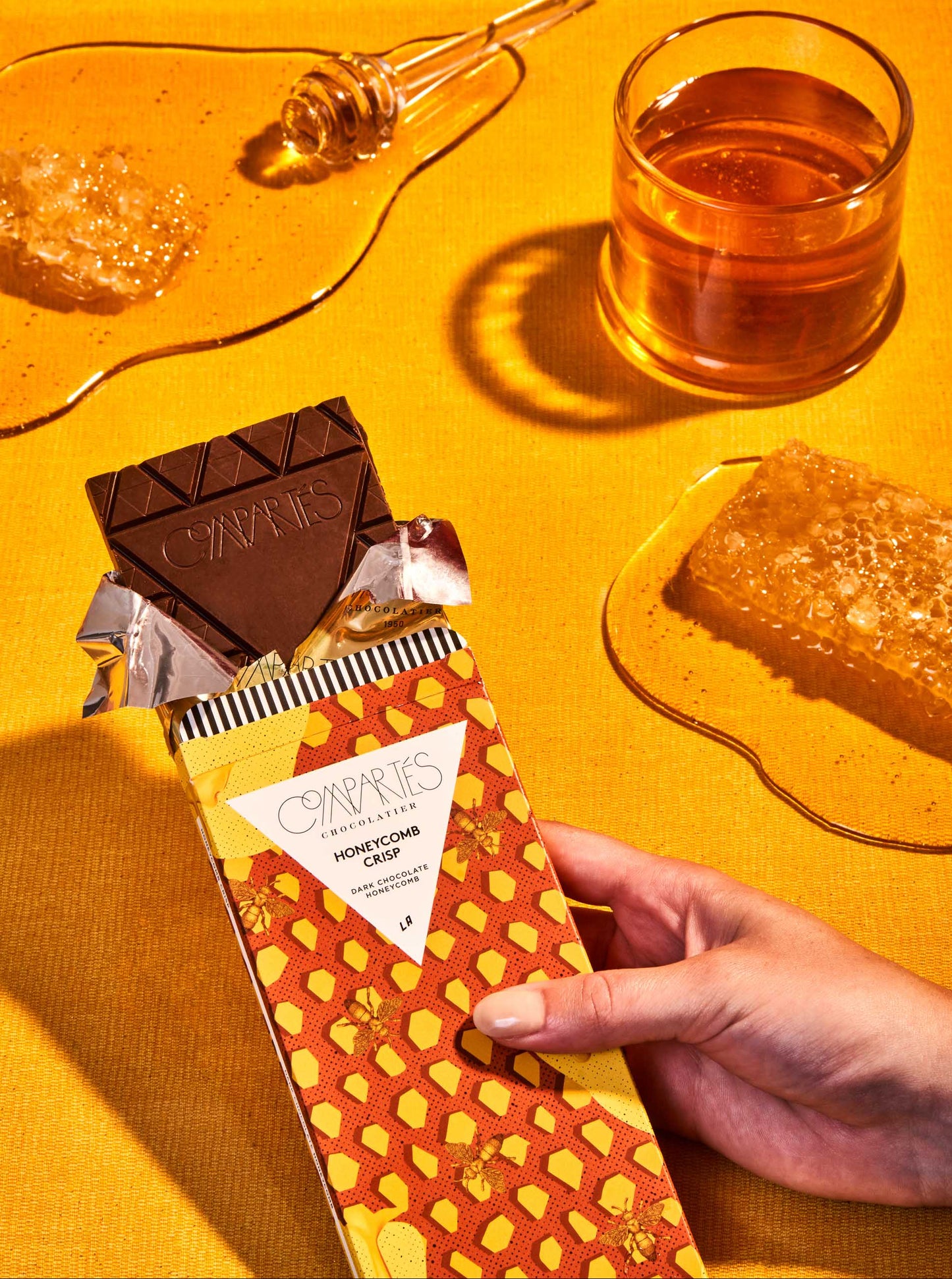A person holding a Honeycomb Crisp Gourmet Dark Chocolate Bar with pieces of honeycomb scattered around on a yellow surface.