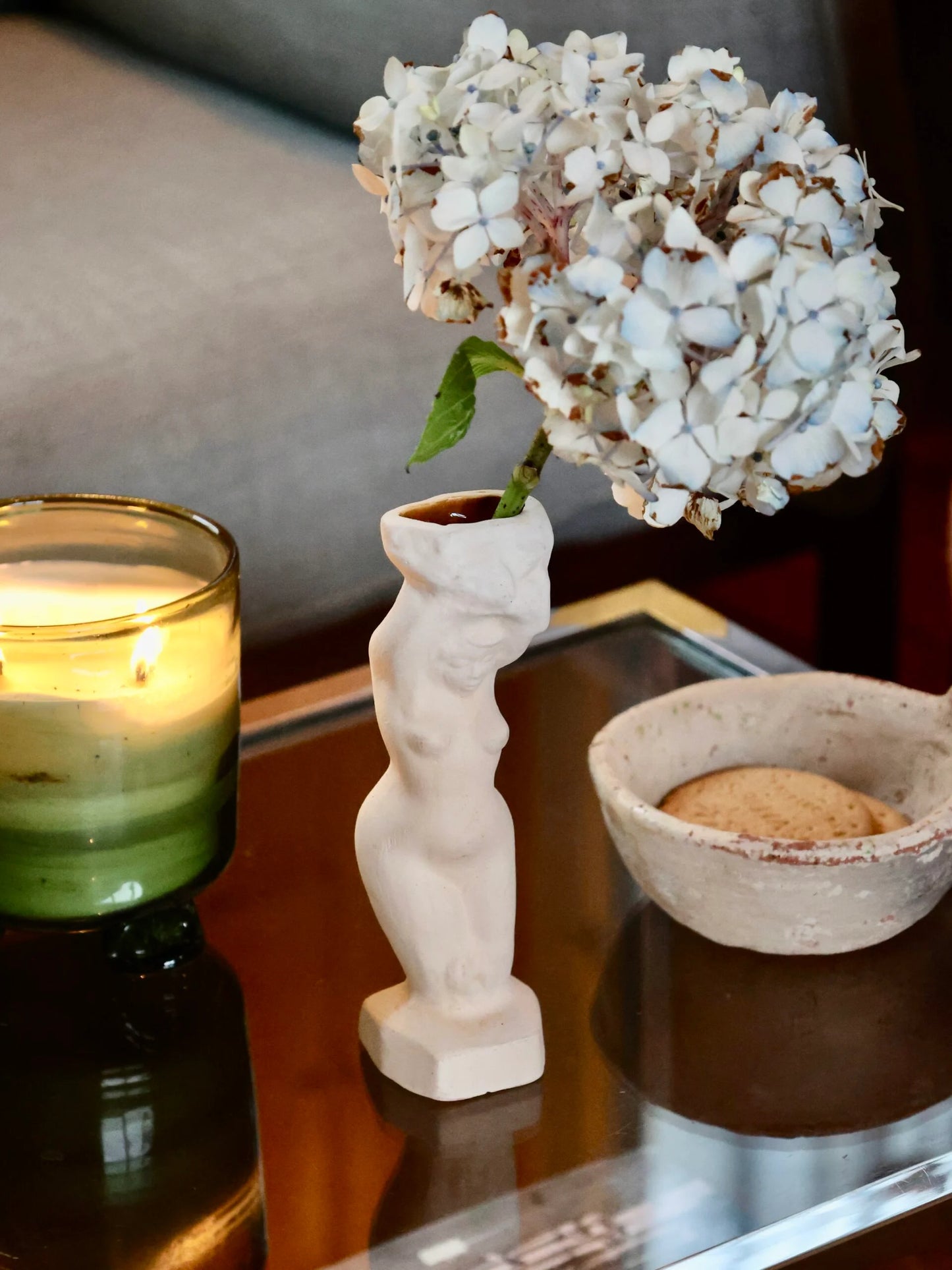Decorative candle, vase resembling a woman’s sculpture with flowers, and bowl on a reflective surface.