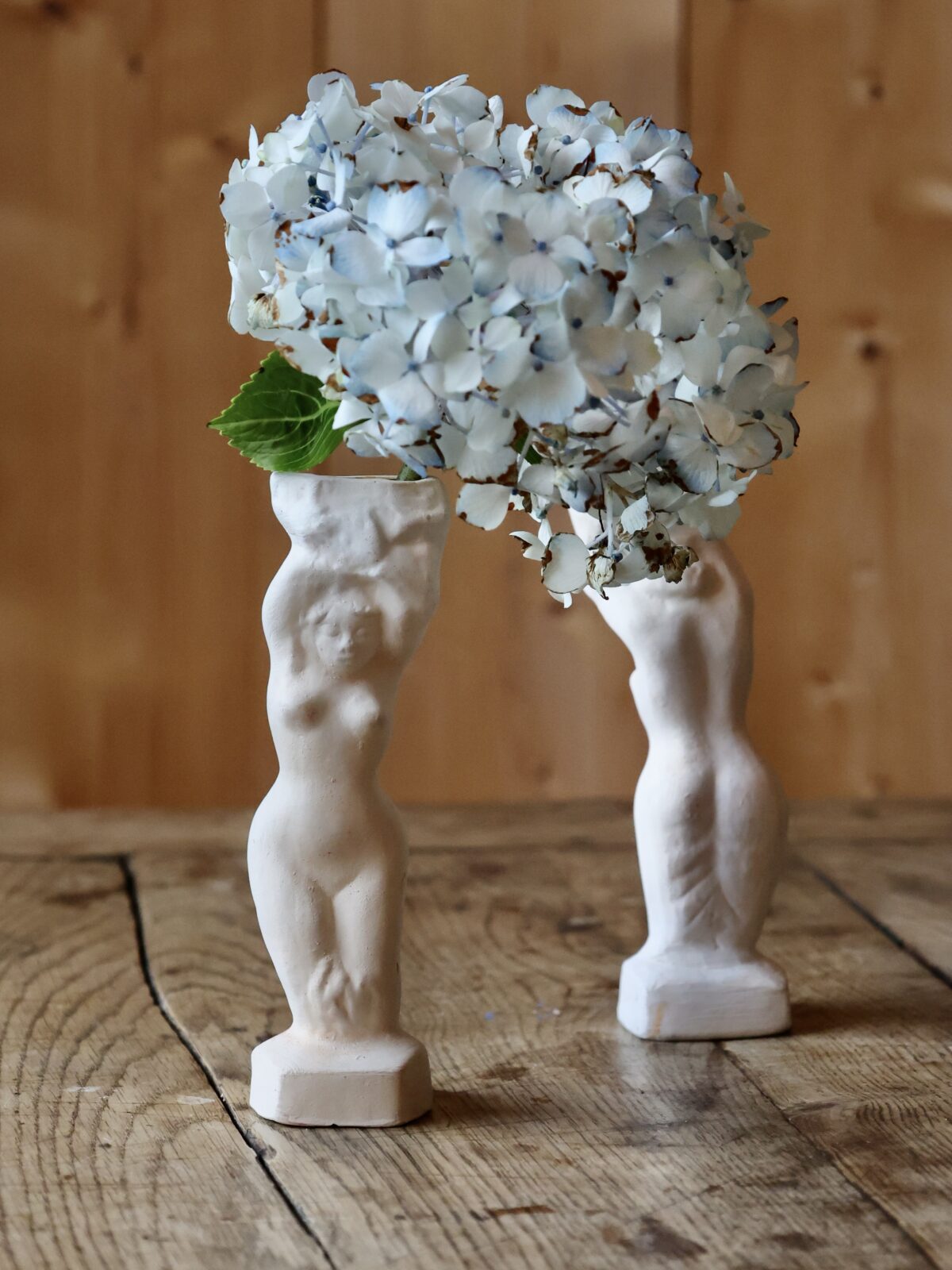 Decorative vase shaped like two standing woman's figures with a bouquet of white flowers on a wooden surface.