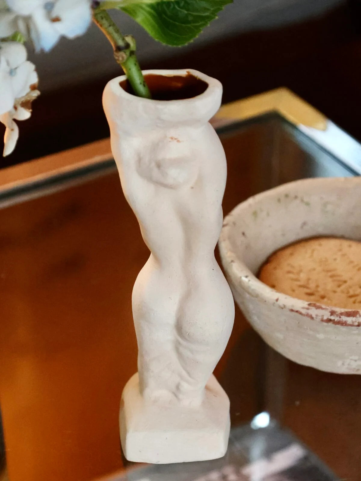 White handmade sculptural vase shaped like a woman with a plant in a pot on a reflective surface.