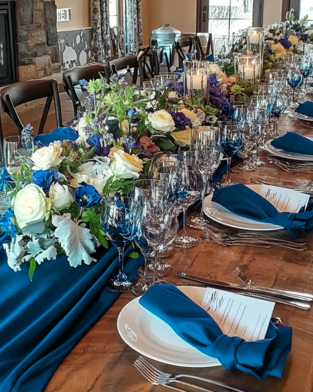Elegant table setting with blue and white floral arrangements, clear glasses, and blue linen napkins.