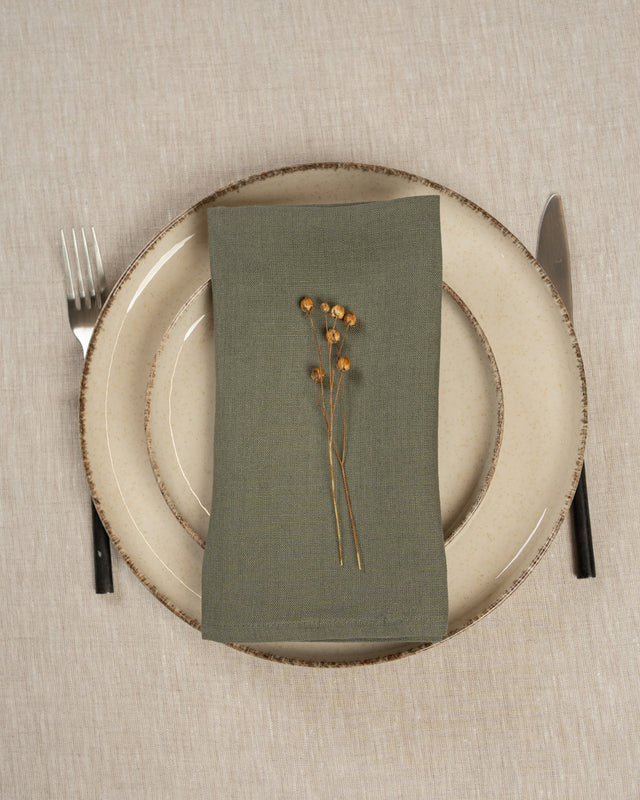 Place setting with a stone green linen napkin, dried flowers, and cutlery on a beige tablecloth.