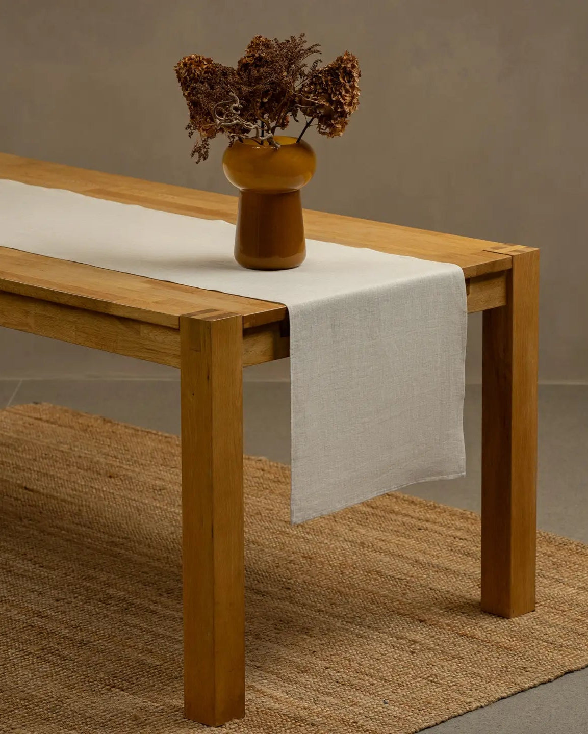 Wooden table with a natural colored runner and a vase with dried flowers on a neutral background.