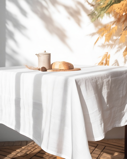 A white 100% linen tablecloth laid out on a dining table, with a small loaf of bread and a pottery item on top, set against a backdrop of natural light and plants.