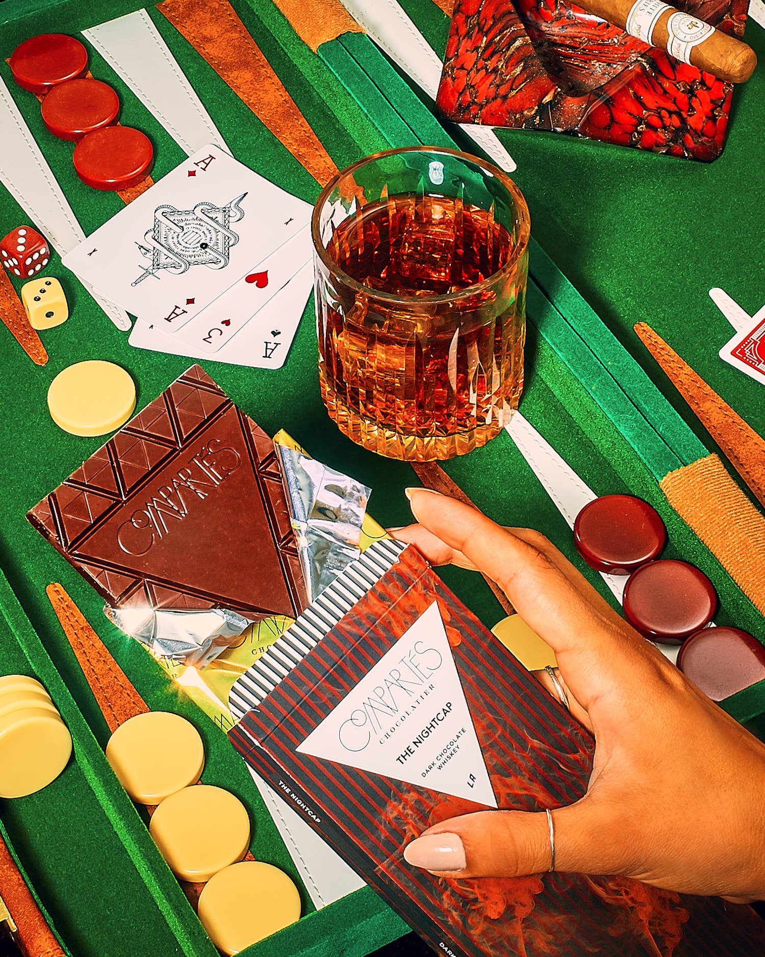 A chocolate bar with a label that reads 'Nightcap Whisky Dark Chocolate Bar' is shown resting on a green surface with a glass of whiskey, a pack of cigarettes, and dice nearby. The background features a poker set and a person's hand reaching for the bar.