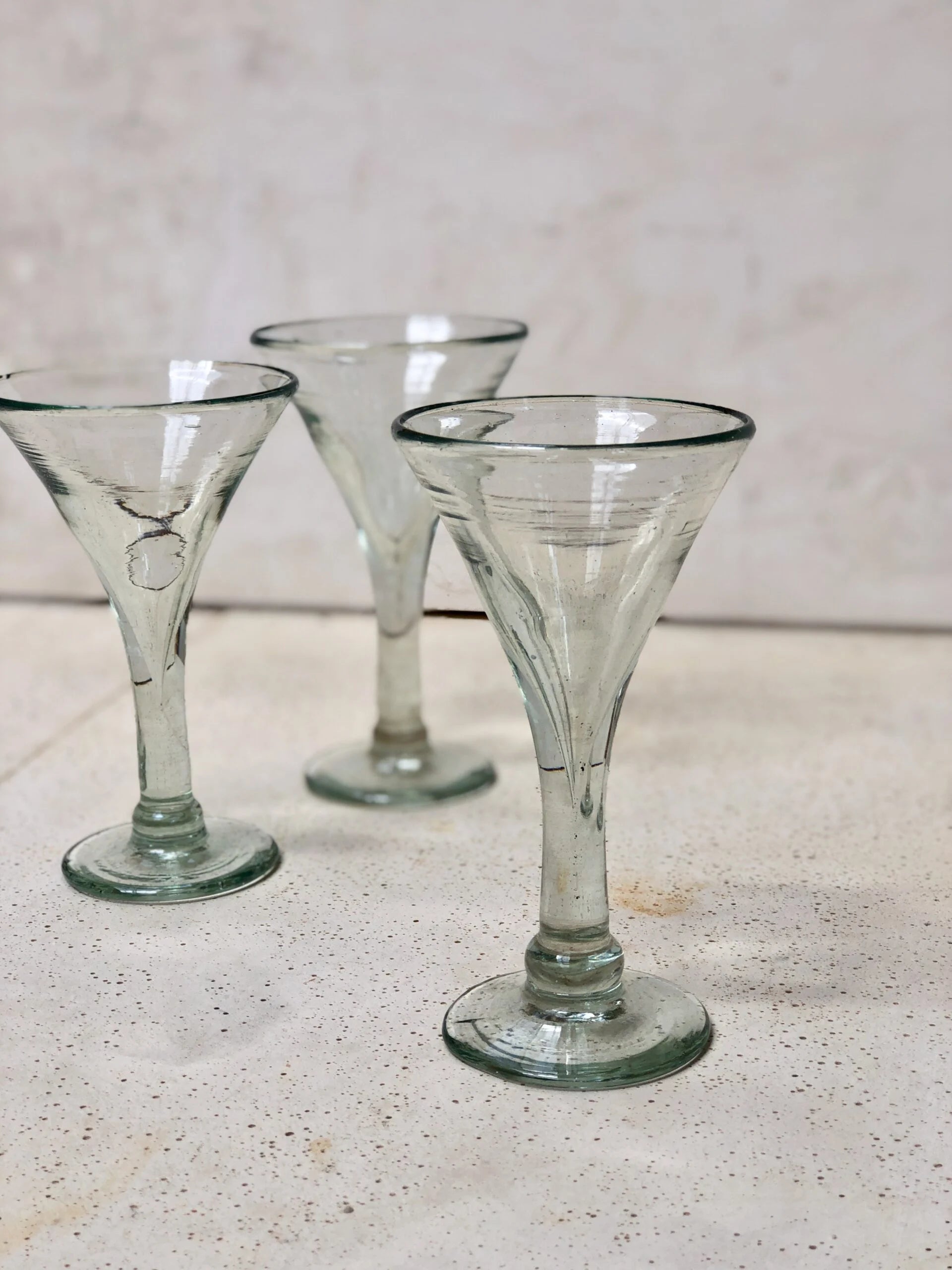 Three elegant vintage-style clear glass cocktail glasses on a light surface with a neutral background. Details are proof that the glasses are mouth-blown.