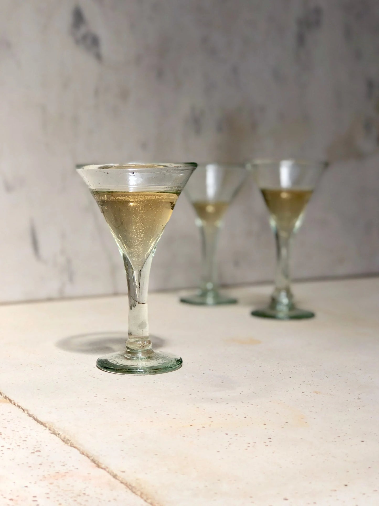 Three artisanal recycled martini glasses with a clear liquid on a textured surface with a neutral background