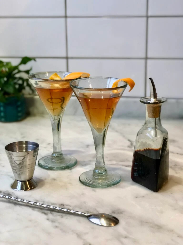 Two La Soufflerie hand-blown martini glasses made from recycled glass, with a cocktail and a bottle of bitters on a marble surface.