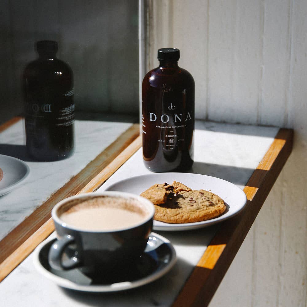 A bottle of Masala Chai Concentrate from the brand Dona, on a coffee shop counter with a cup of chai latte and a cookie