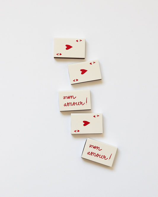 A beige-colored matchbox with the text 'Mon Amour' and a heart symbol printed in a red color on the front, symbolizing  an ace of hearts card.
