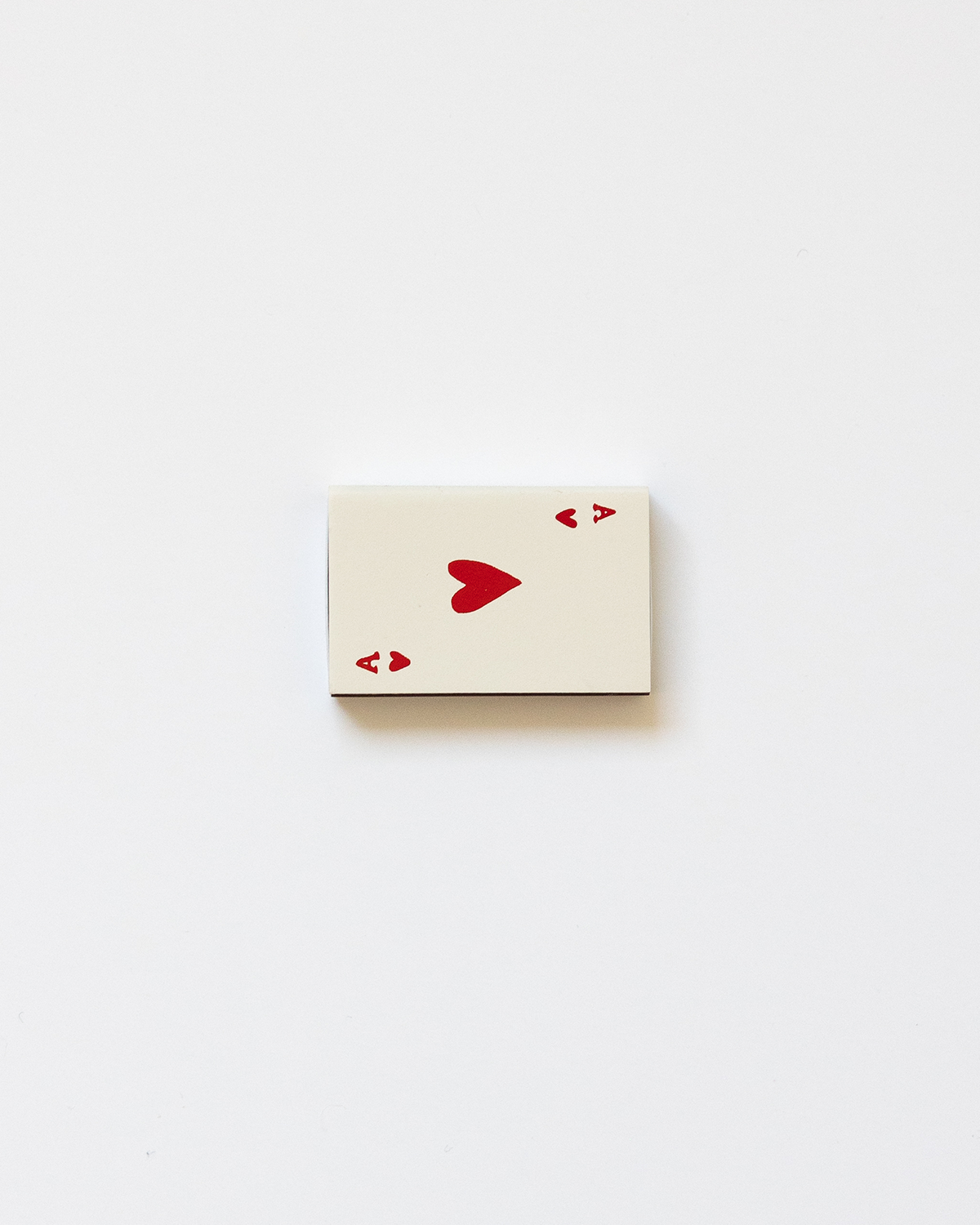 A beige-colored matchbox with a heart symbol printed in a red color on the front, symbolizing an ace of hearts card.