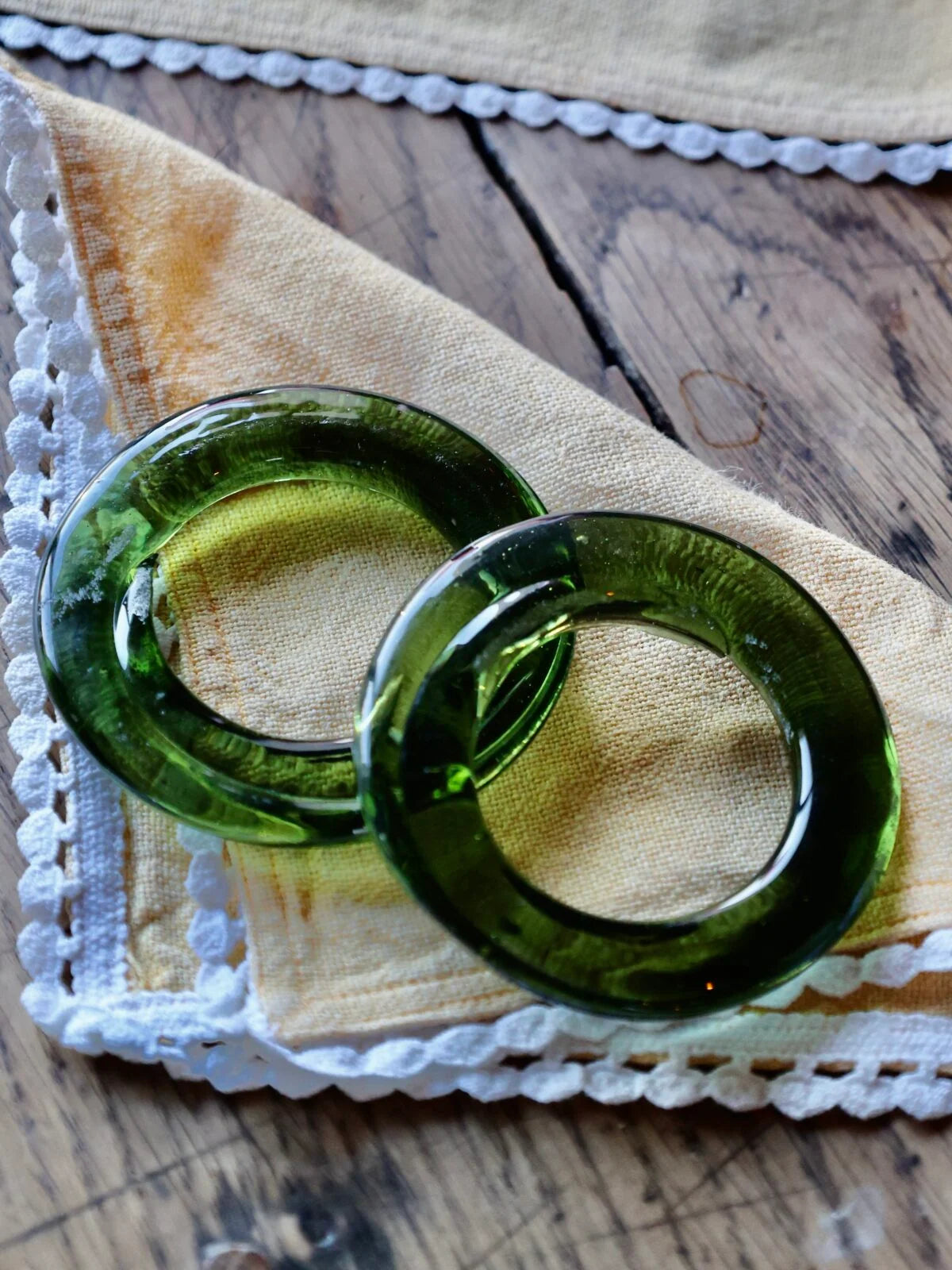 Two smoky green glass napkin rings on a textured fabric surface with lace trim.