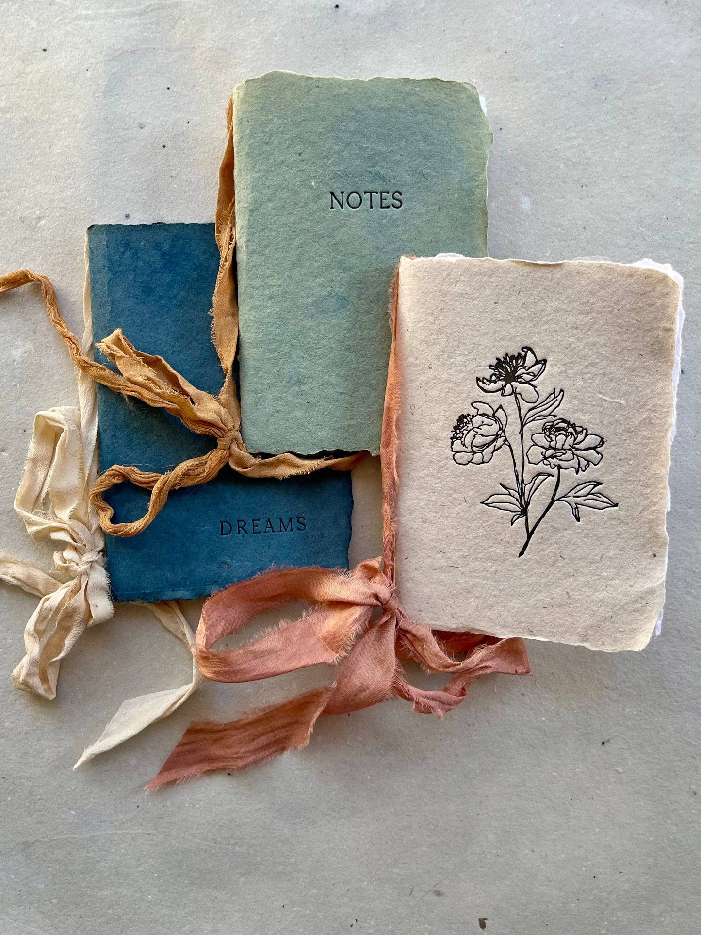 Three handmade notebooks with silk ribbons and botanical-dyed covers, two labeled 'NOTES' and 'DREAMS', and one with a floral drawing.