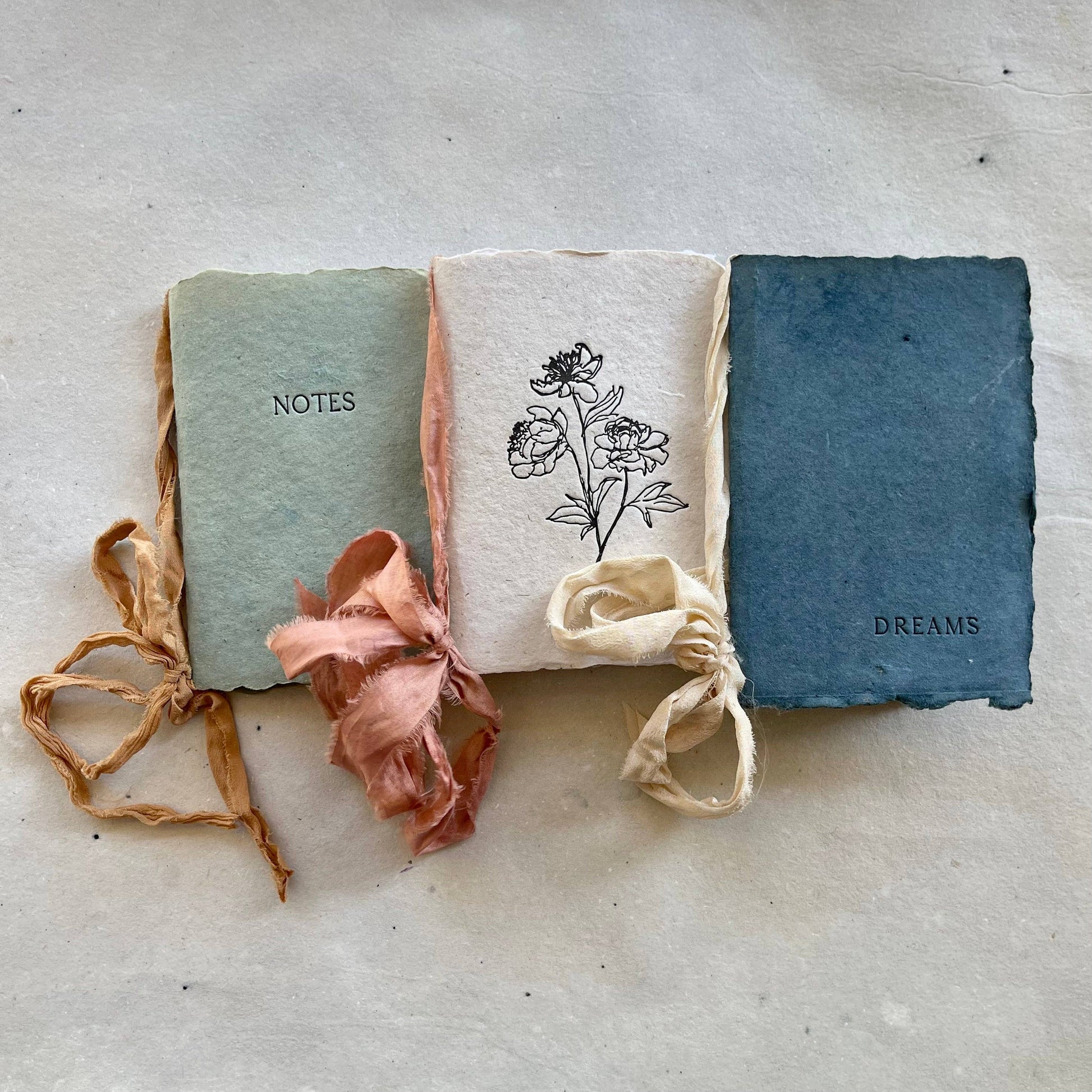 Three handmade notebooks with silk ribbons and botanical-dyed covers, two labeled 'NOTES' and 'DREAMS', and one with a floral drawing.