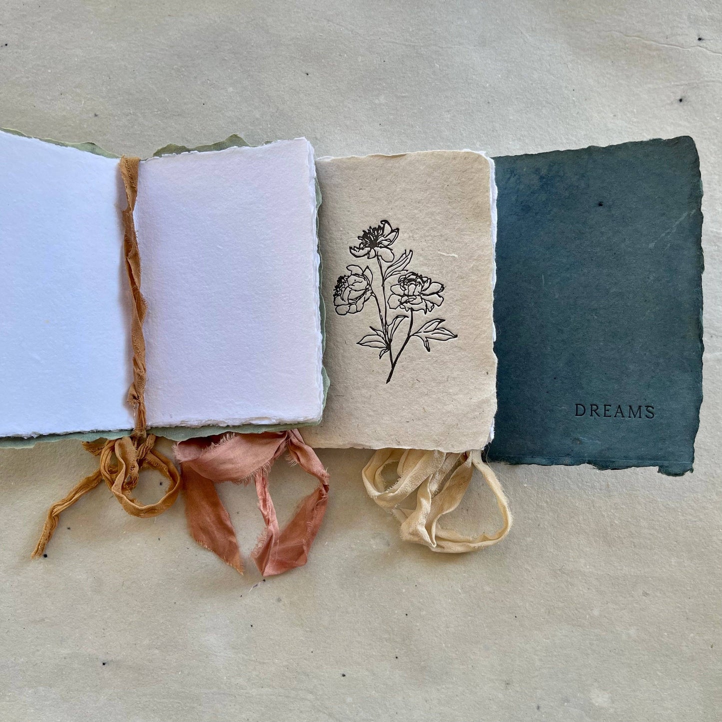 Three handmade notebooks with silk ribbons and botanical-dyed covers, two labeled 'NOTES' and 'DREAMS', and one with a floral drawing. One notebook is open, showing that the interior pages are white.