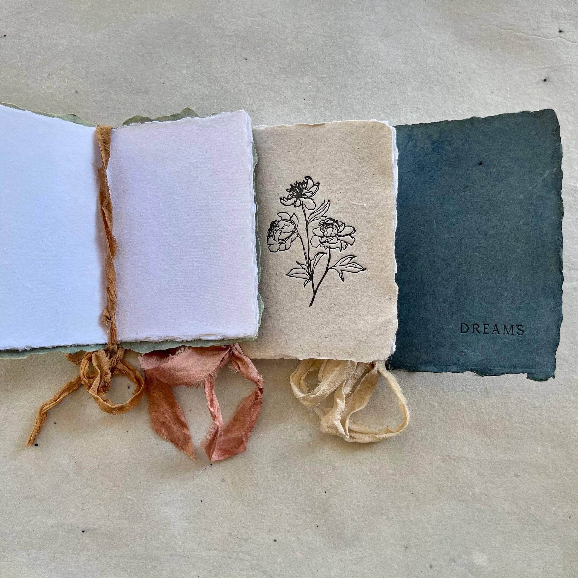 Three handmade notebooks with silk ribbons and botanical-dyed covers, two labeled 'NOTES' and 'DREAMS', and one with a floral drawing. One notebook is open, showing that the interior pages are white.
