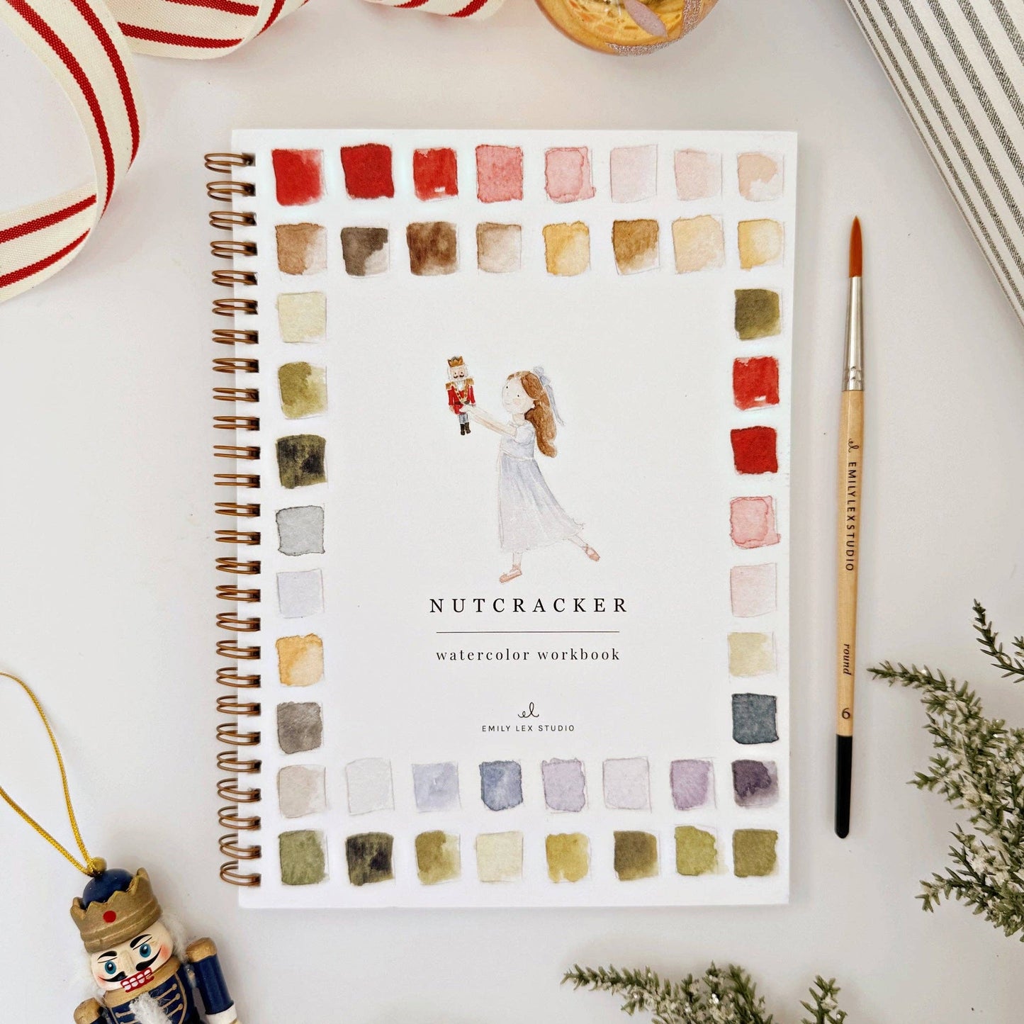Watercolor workbook titled 'Nutcracker' with color swatches, and Christmas-themed decor around it, on a white surface.