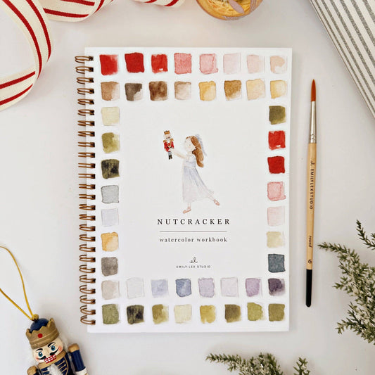 Watercolor workbook titled 'Nutcracker' with color swatches, and Christmas-themed decor around it, on a white surface.
