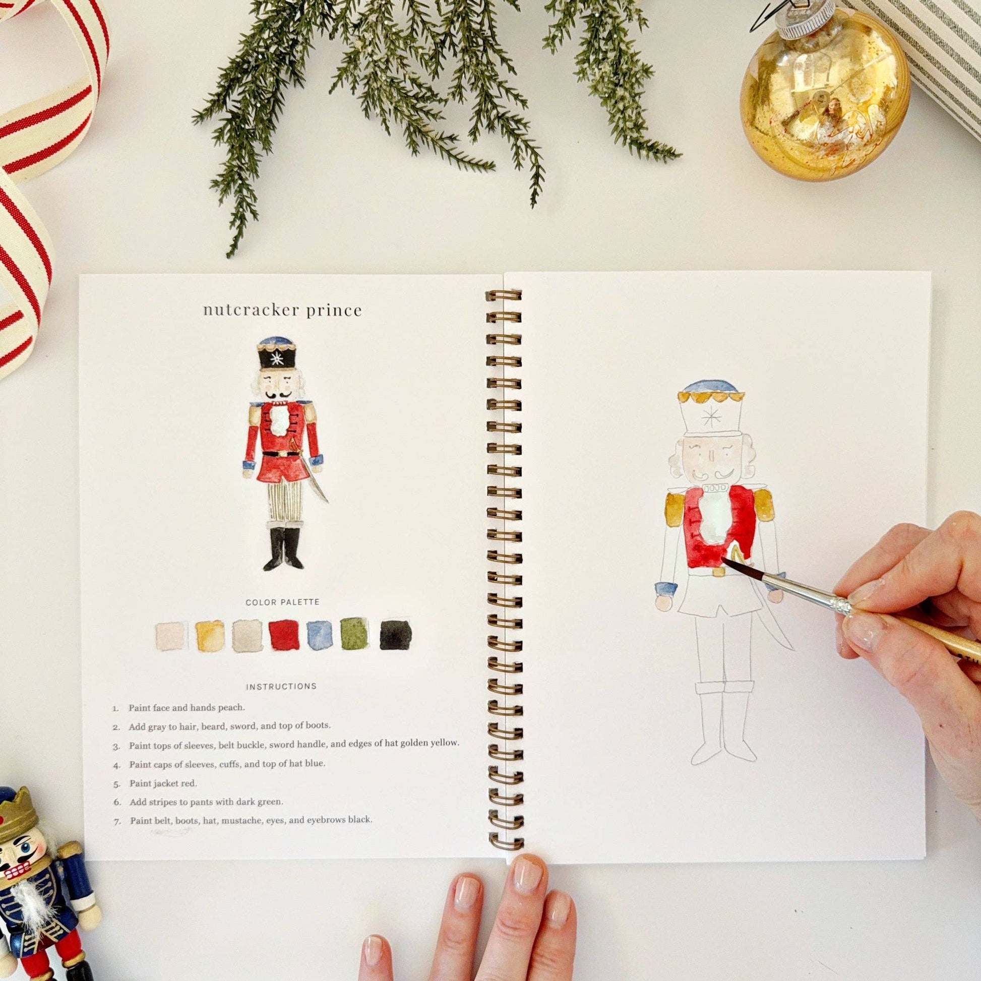 Open watercolor workbook with a nutcracker illustration, color chart and step-by-step instructions, surrounded by Christmas decorations.
