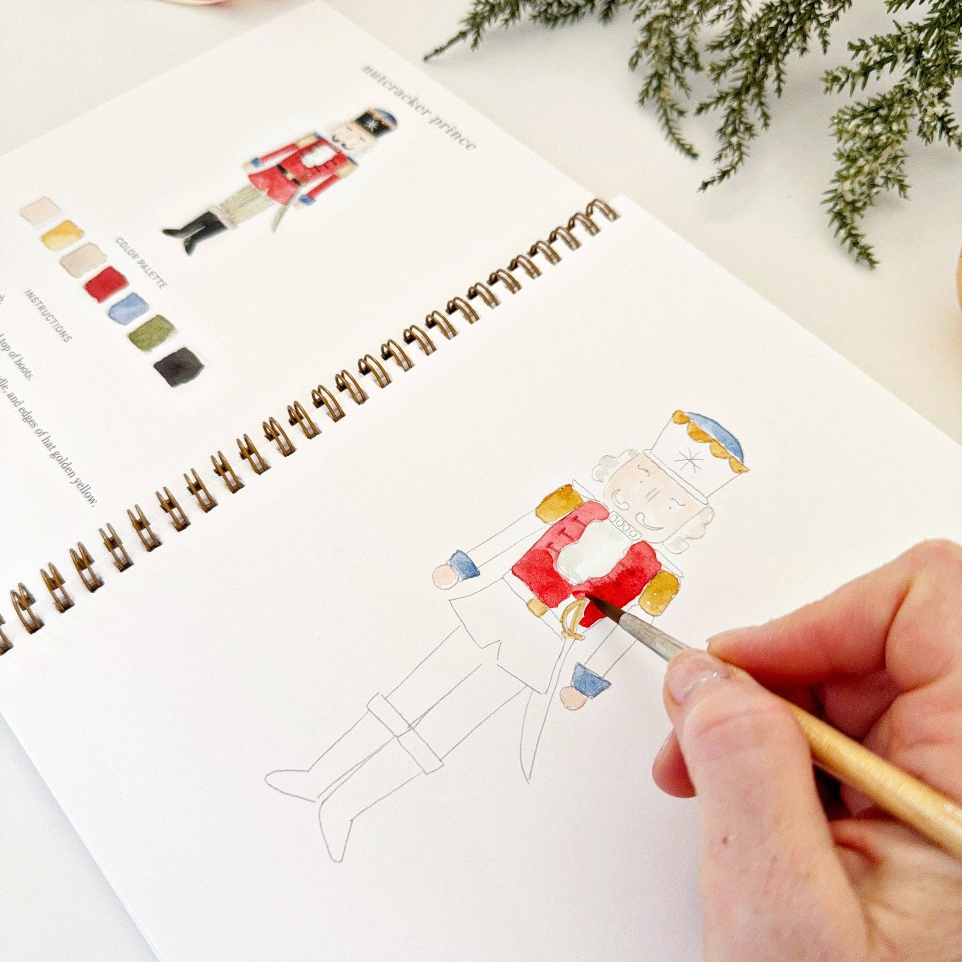 Person coloring a nutcracker illustration in a watercolor sketchbook with a color swatch guide.