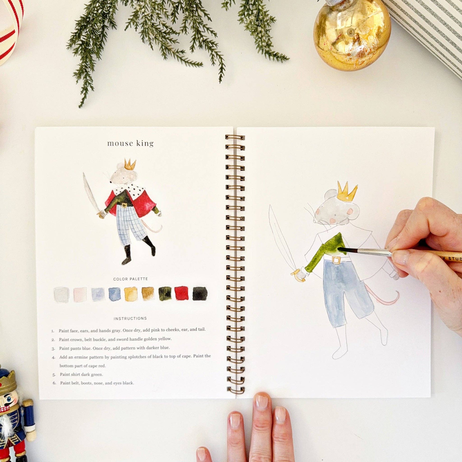 Person coloring a mouse king illustration in a watercolor coloring book with a color palette on a white surface, surrounded by Christmas decorations.