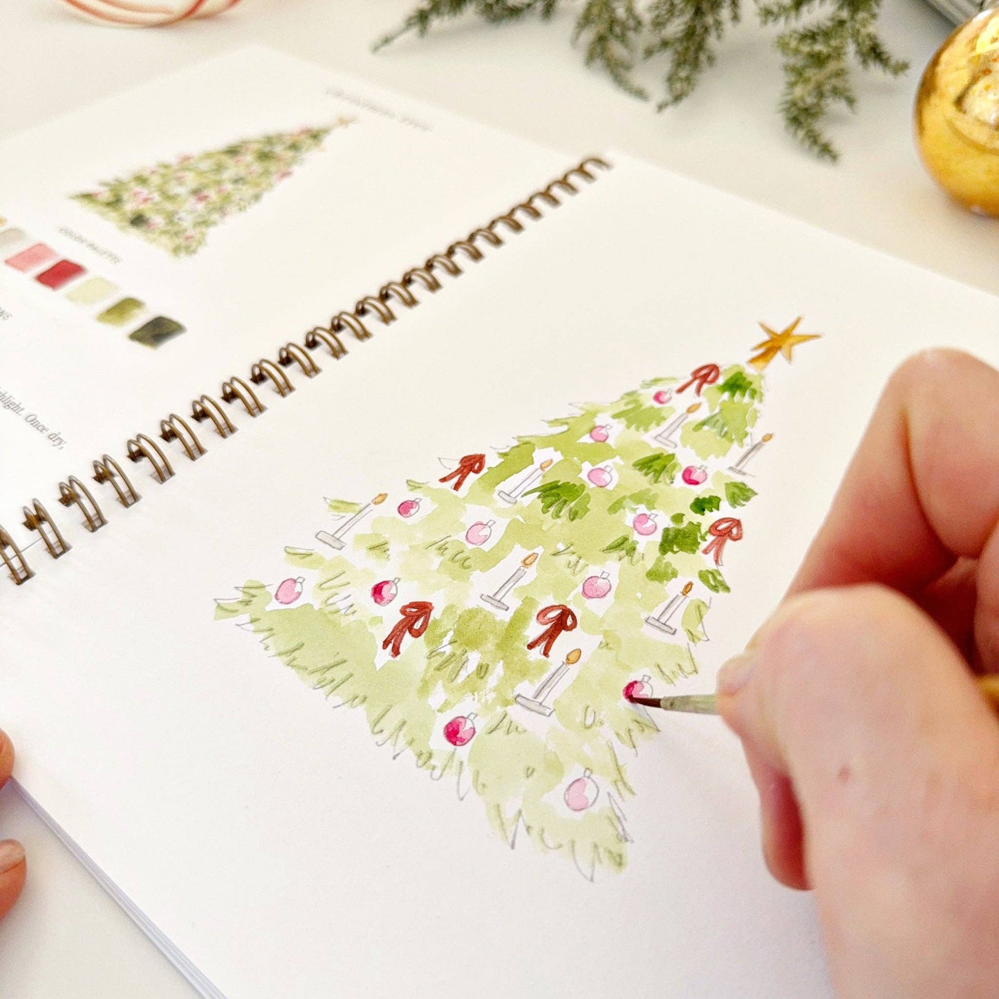 Person drawing a Christmas tree on a spiral-bound watercolor workbook with a brush.