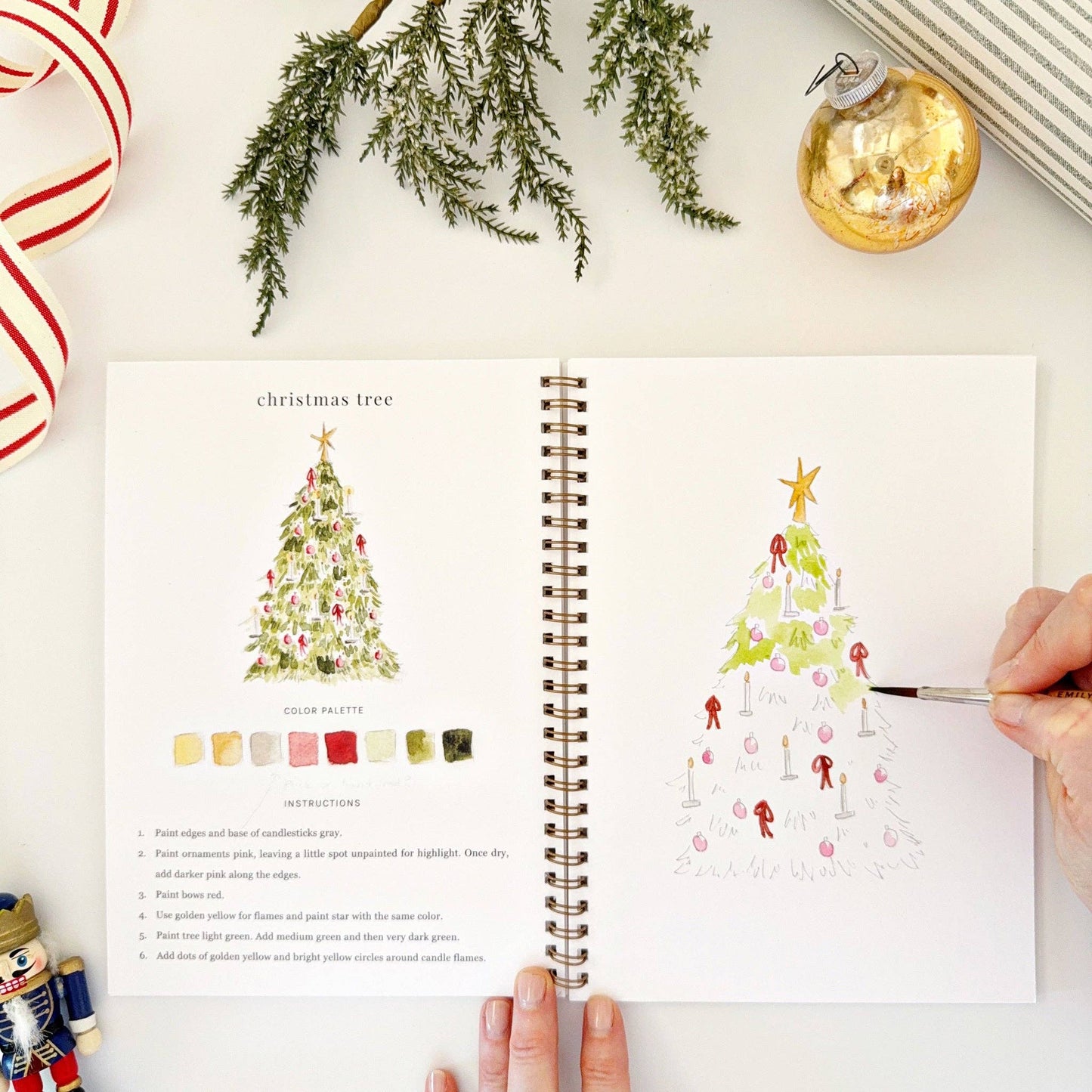 Open watercolor book with Christmas tree illustrations and color palette on a white surface with festive decorations.