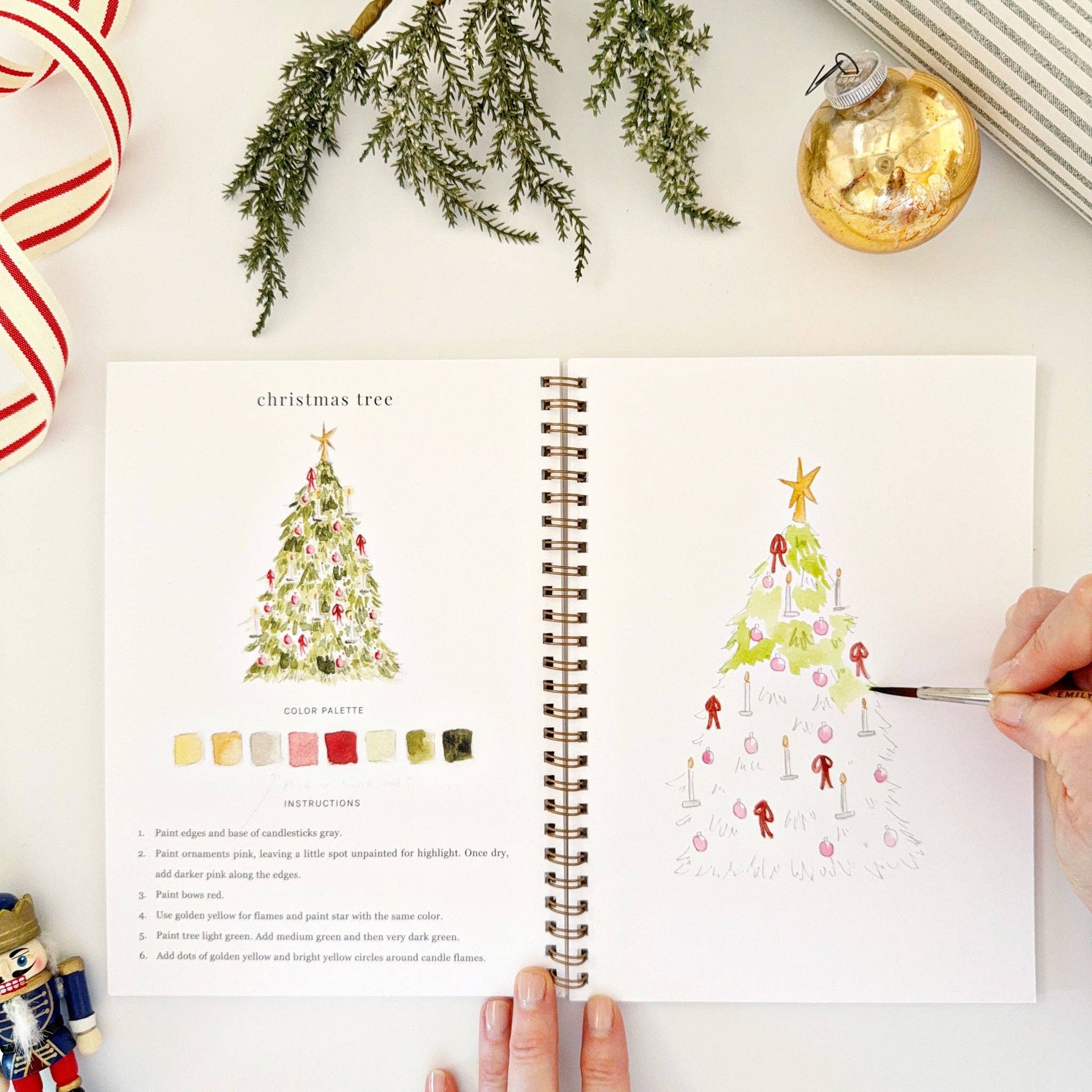 Open watercolor book with Christmas tree illustrations and color palette on a white surface with festive decorations.