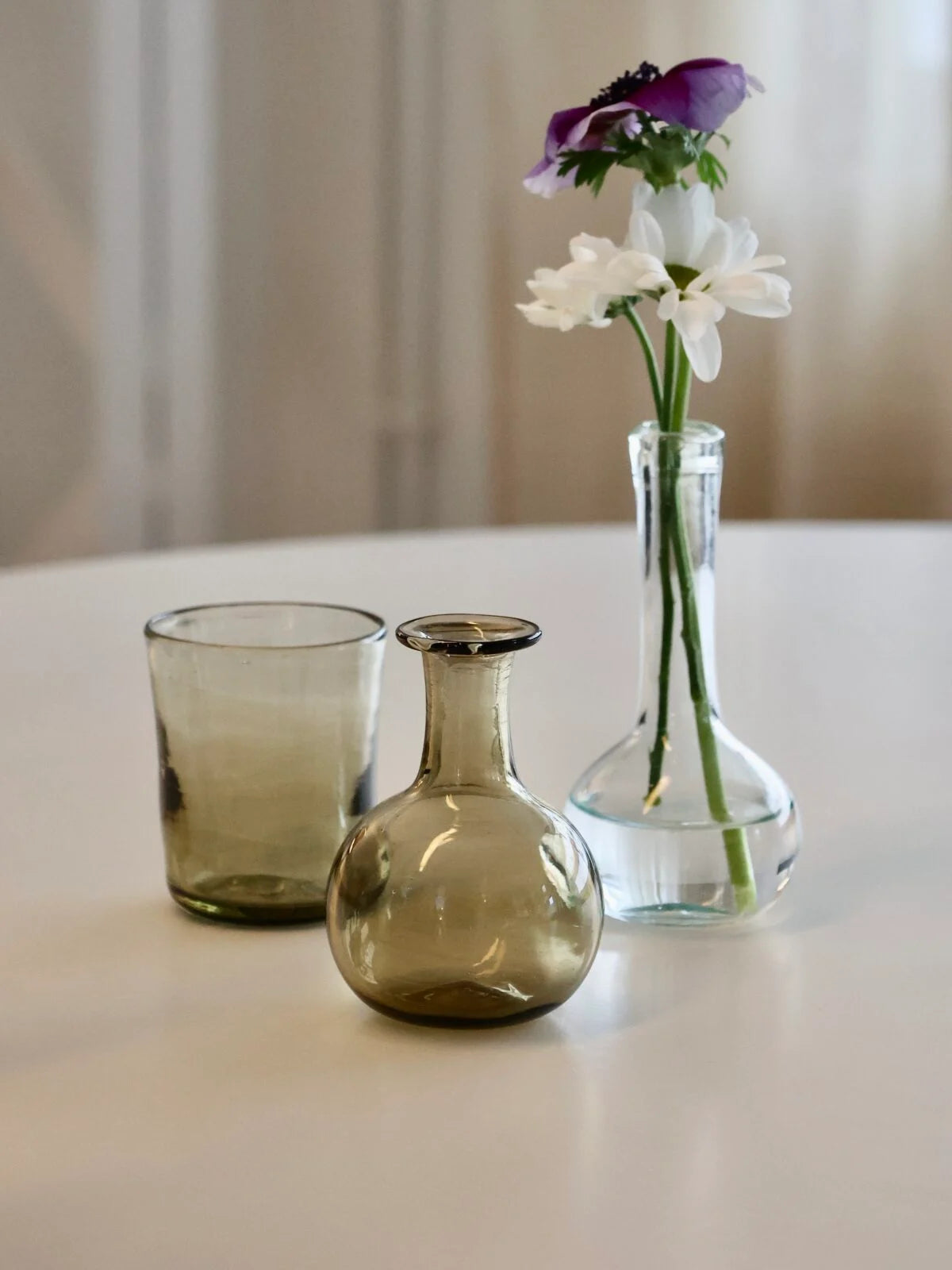 Two handmade glass vases on a white surface with a third vase containing flowers.