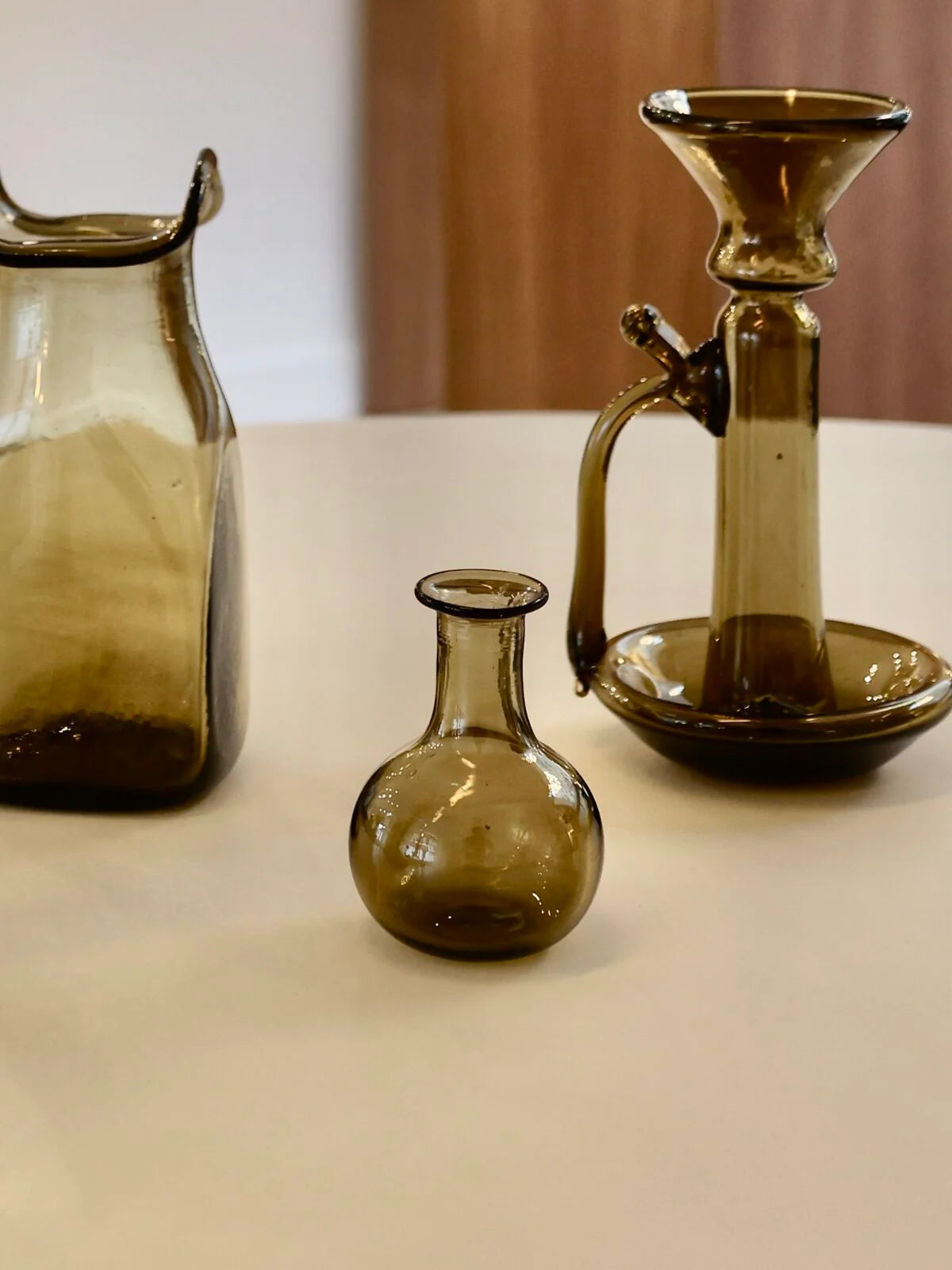 Three brown glass items on a beige surface with a neutral background. The one in the center is a small smoky glass vase with rounded belly and flared neck.