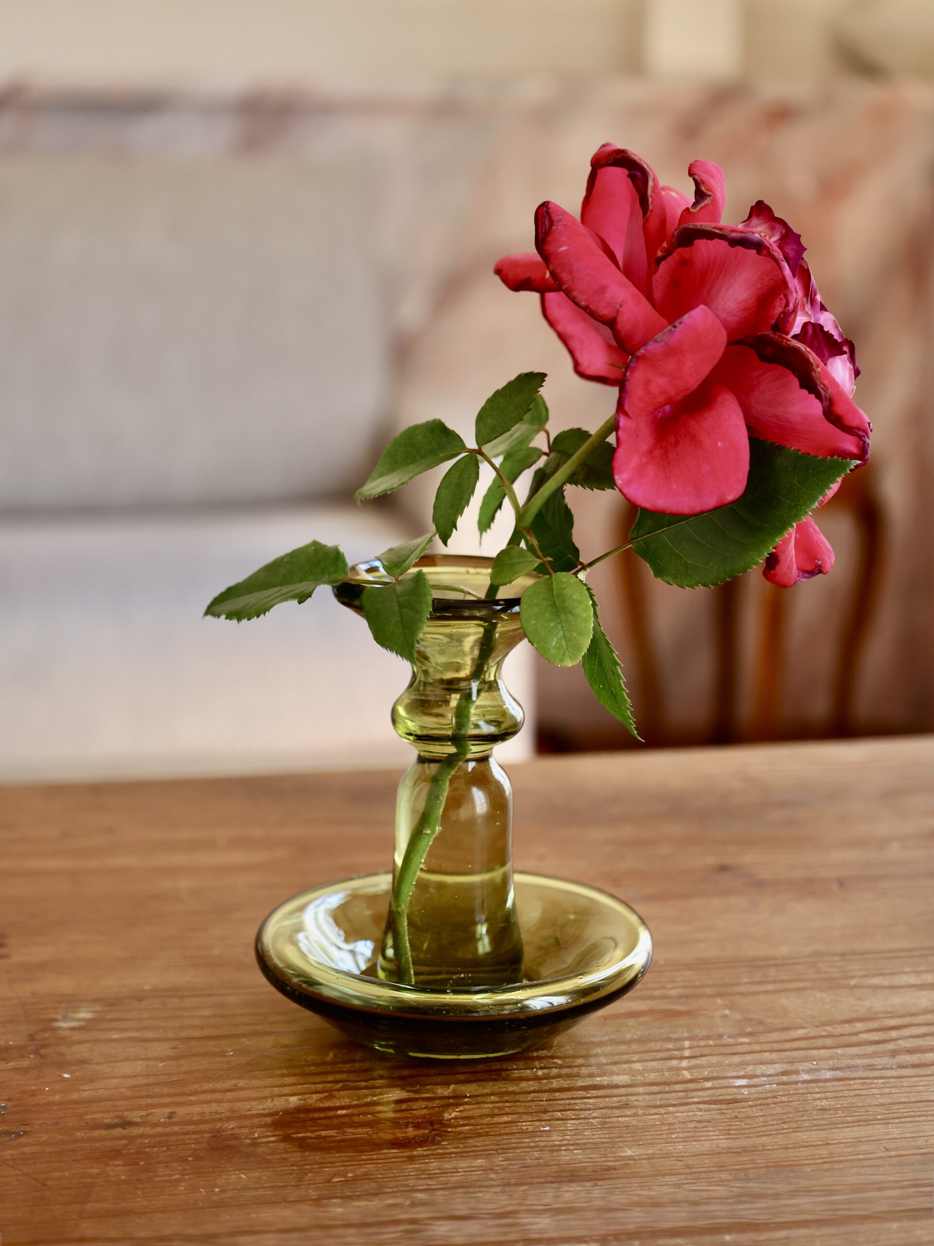 Pink rose in a small handmade glass vase on a wooden surface with a blurred background