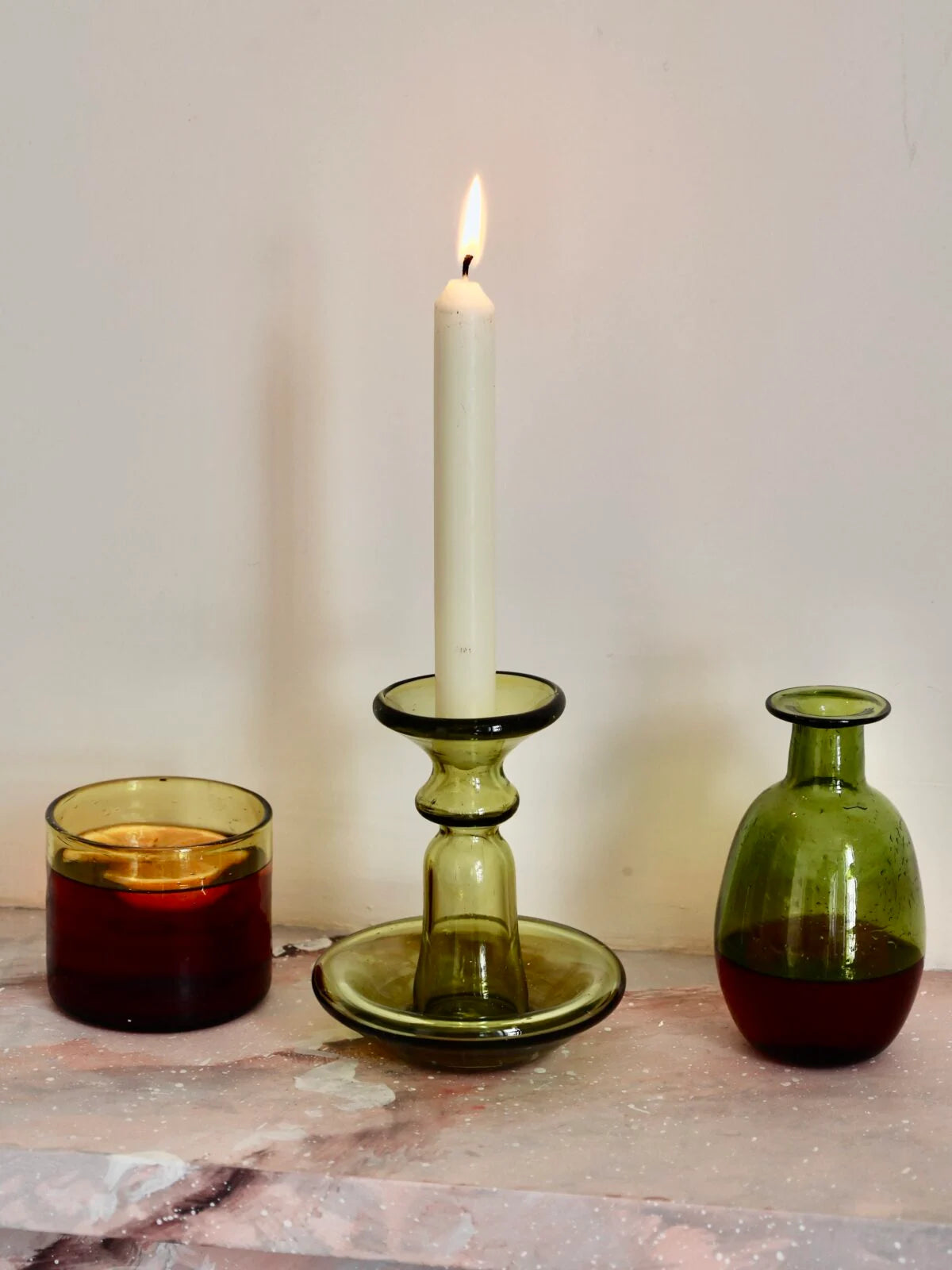 Artisanal green glass candle holder with a lit candle and two green glass containers on a textured surface