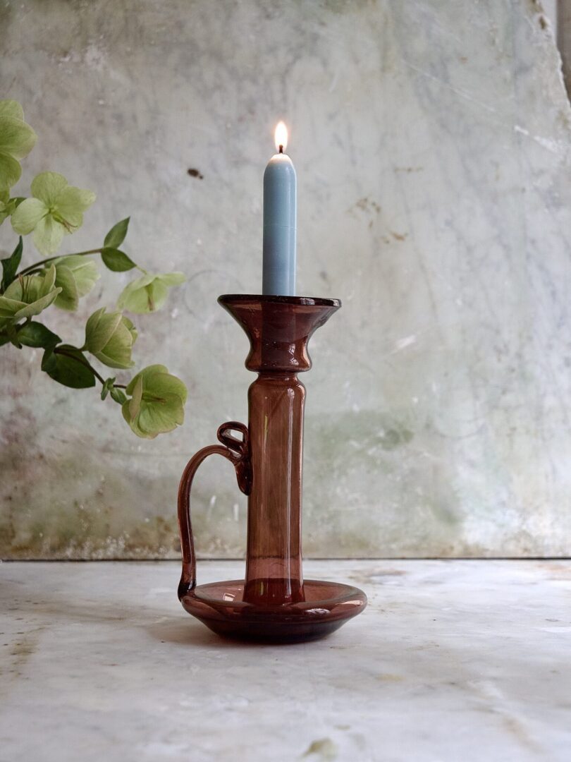 Brown and raspberry-hued candle holder with a lit candle on a textured surface with greenery.