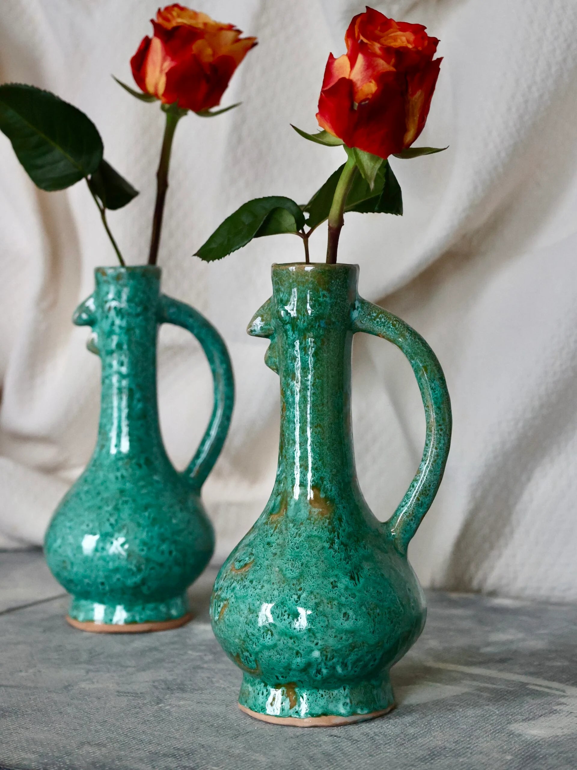 Two handmade turquoise ceramic chicken-shaped vases holding red roses on a neutral background.