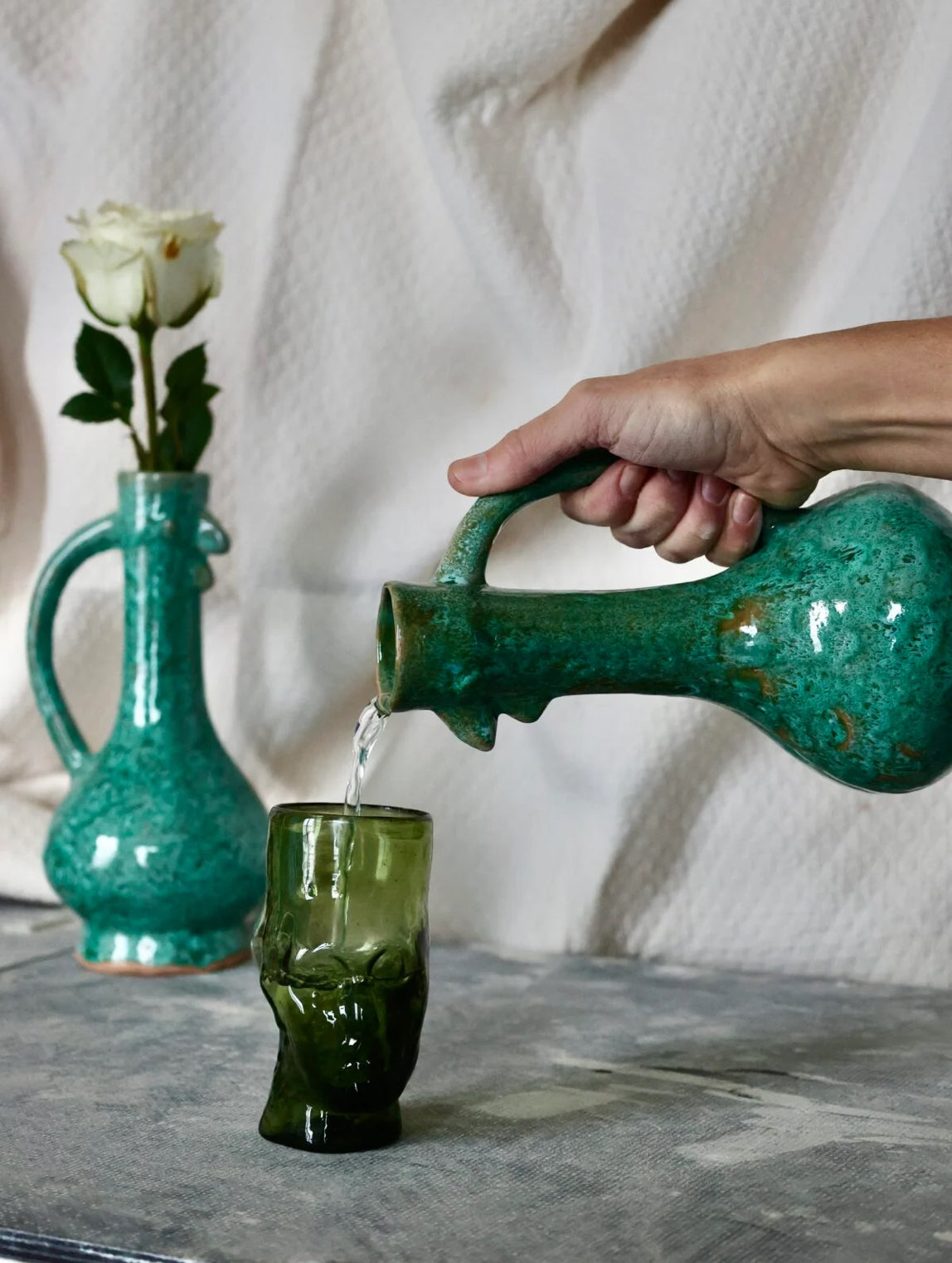 Person pouring water from a turquoise ceramic chicken-shaped pitcher into a glass on a marble surface.