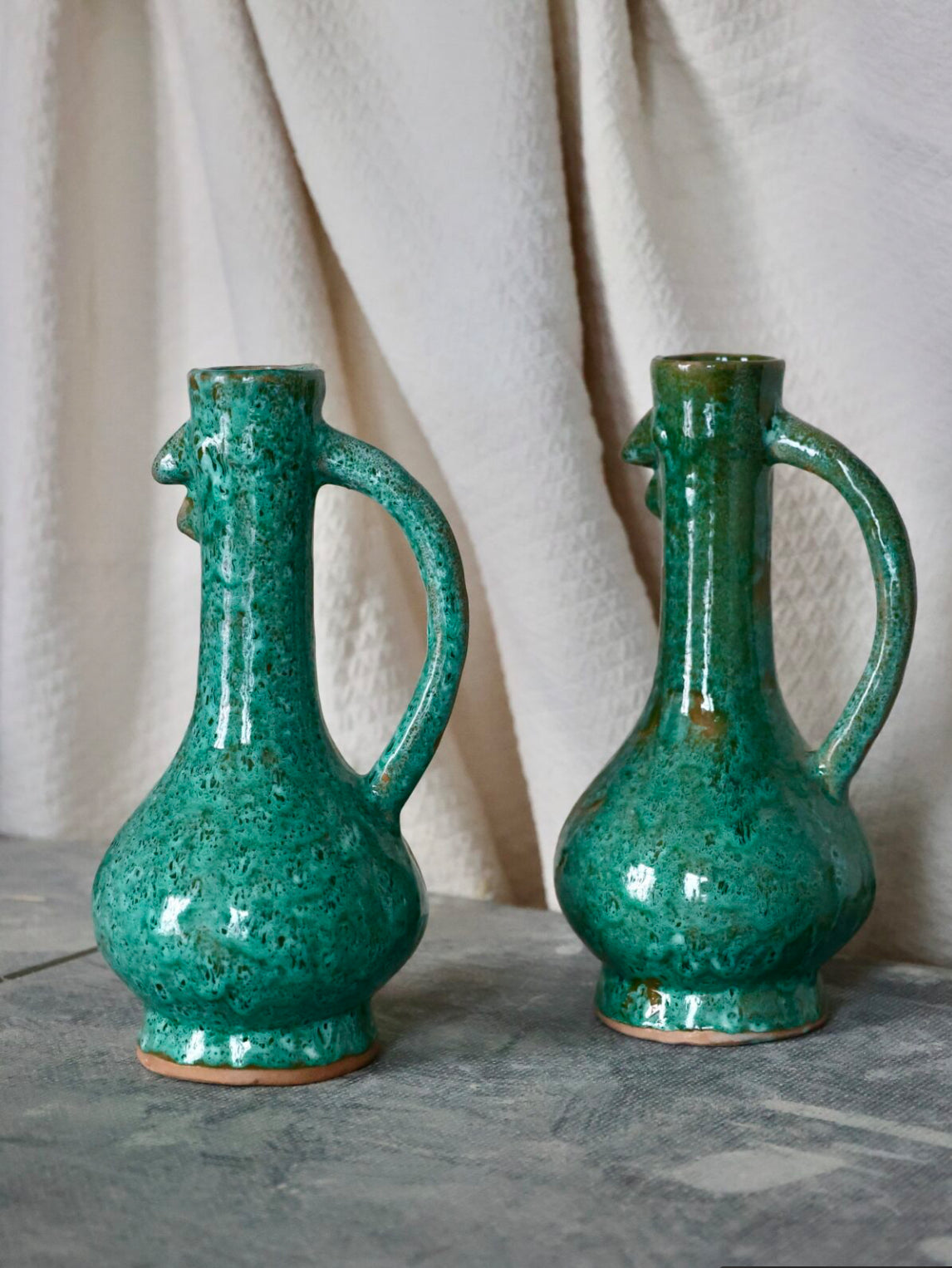 Two turquoise countryside-inspired decorative ceramic vases with handles on a stone surface and light fabric background