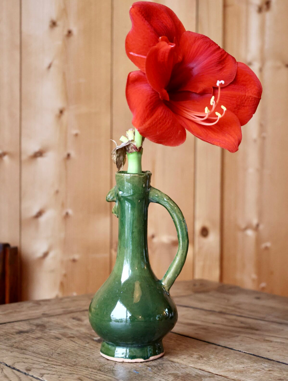 Red flower in a green ceramic chicken-shaped vase on a wooden surface with a wooden panel background
