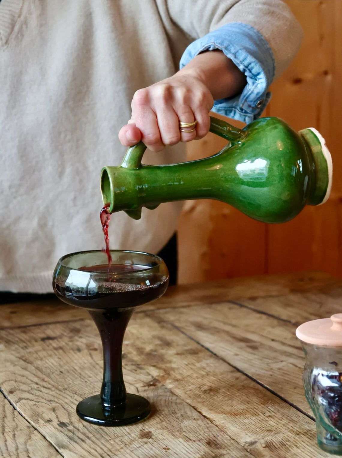 Person pouring red liquid from a green ceramic chicken-shaped carafe into a glass on a wooden surface.