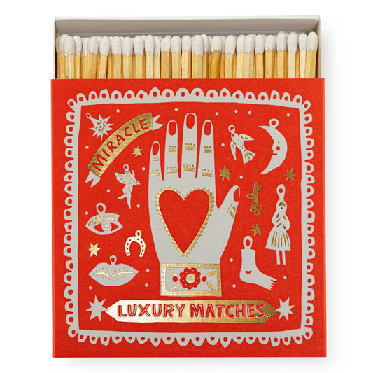 The archivist gallery's signature square matchbox featuring the illustration of a hand with a heart in the middle surrounded by an array of different charms and gold foil detailing with the words 'Miracle Luxury Matches.'