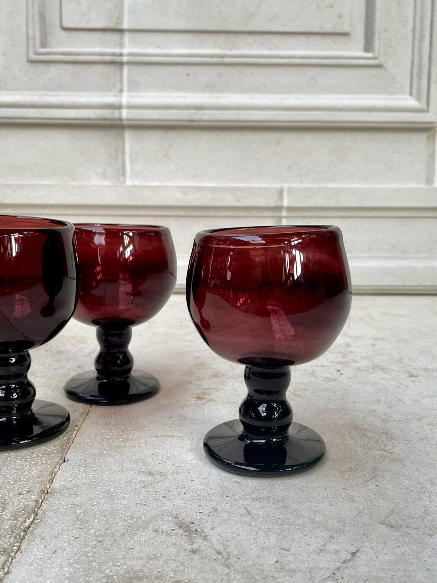 Three red mouth-blown wine glasses on a marble surface with a white marble wall in the background.