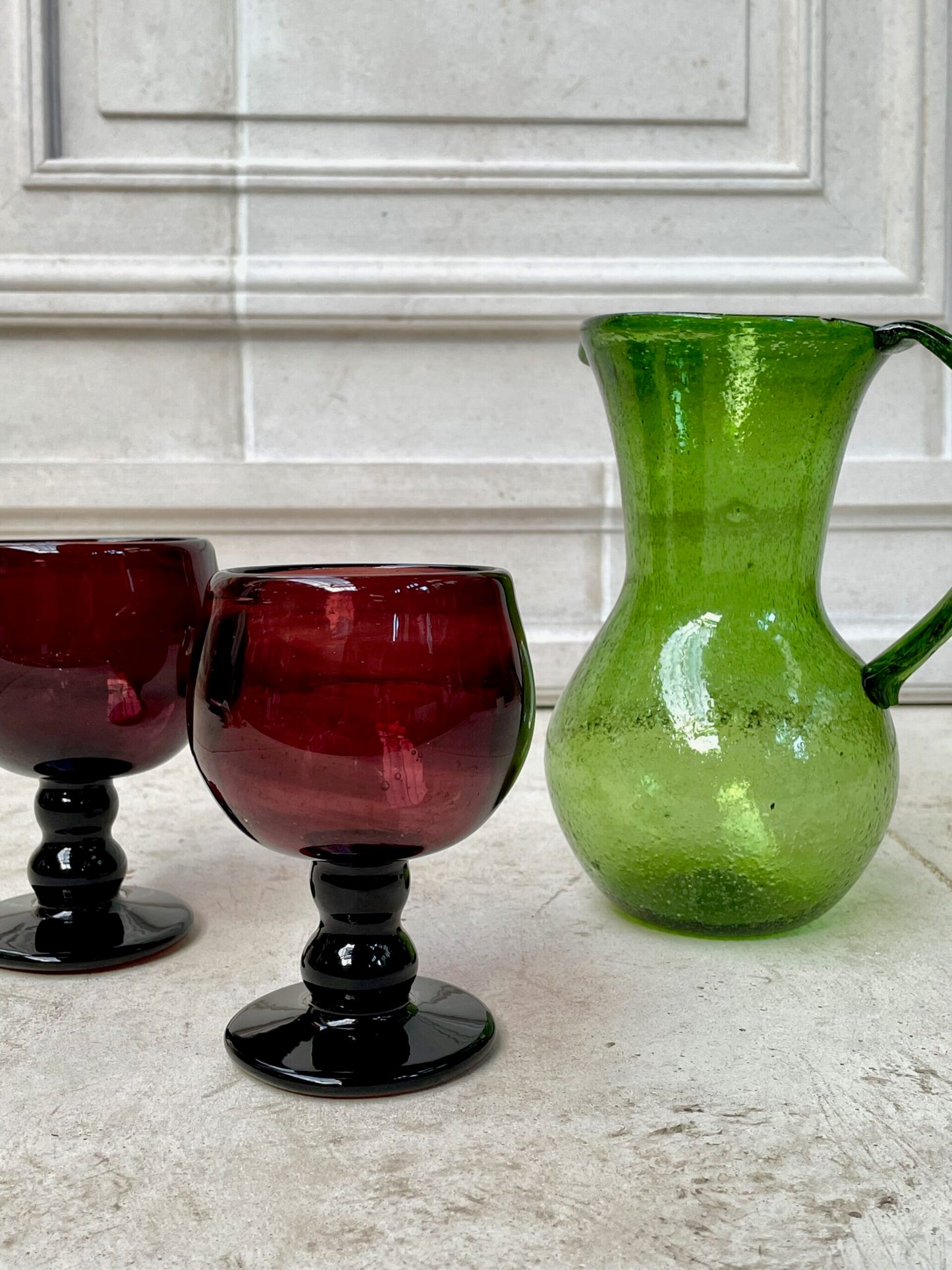 Two sustainable French-style red wine glasses and a green glass pitcher on a countertop.