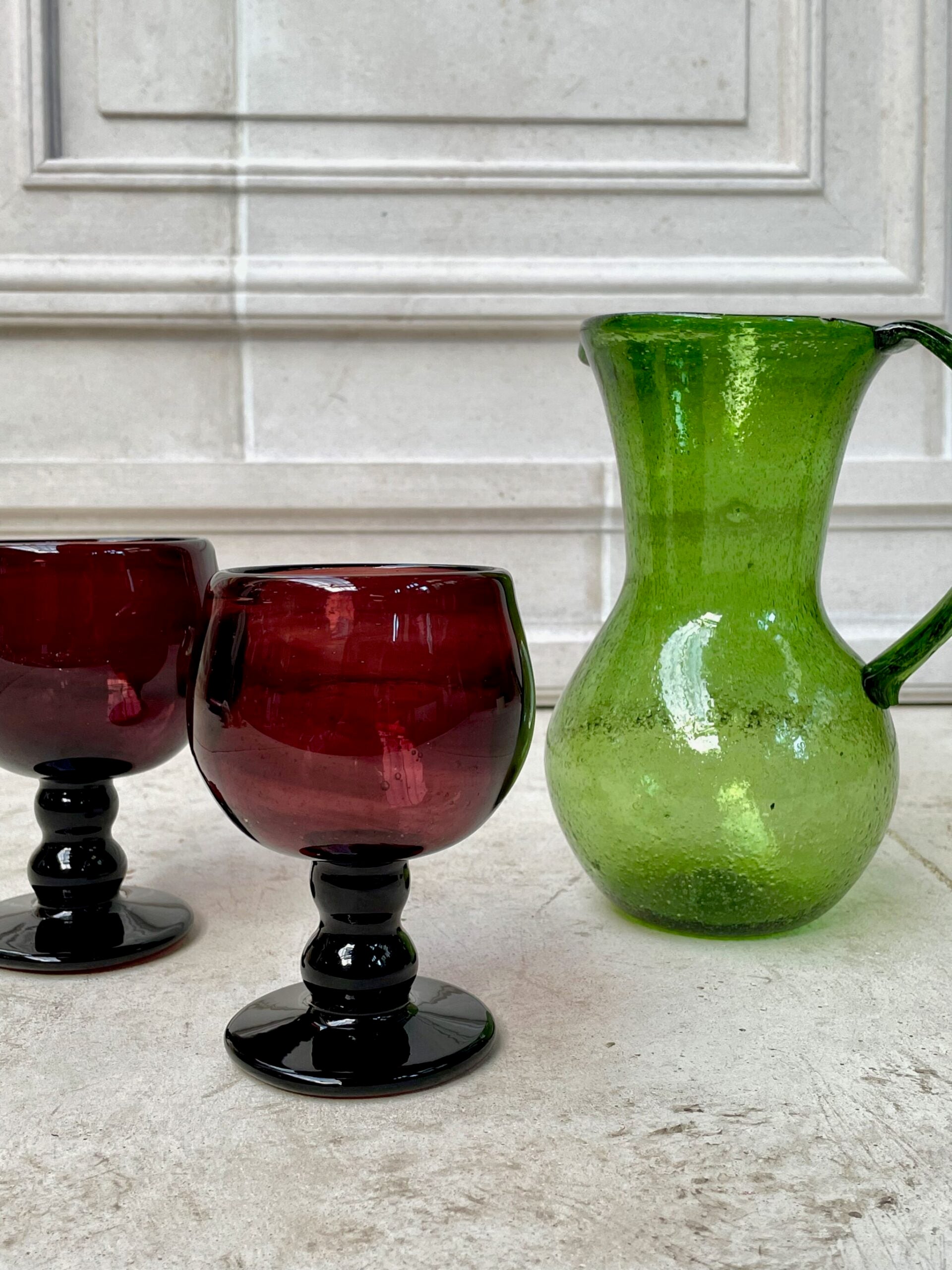 Two sustainable French-style red wine glasses and a green glass pitcher on a countertop.