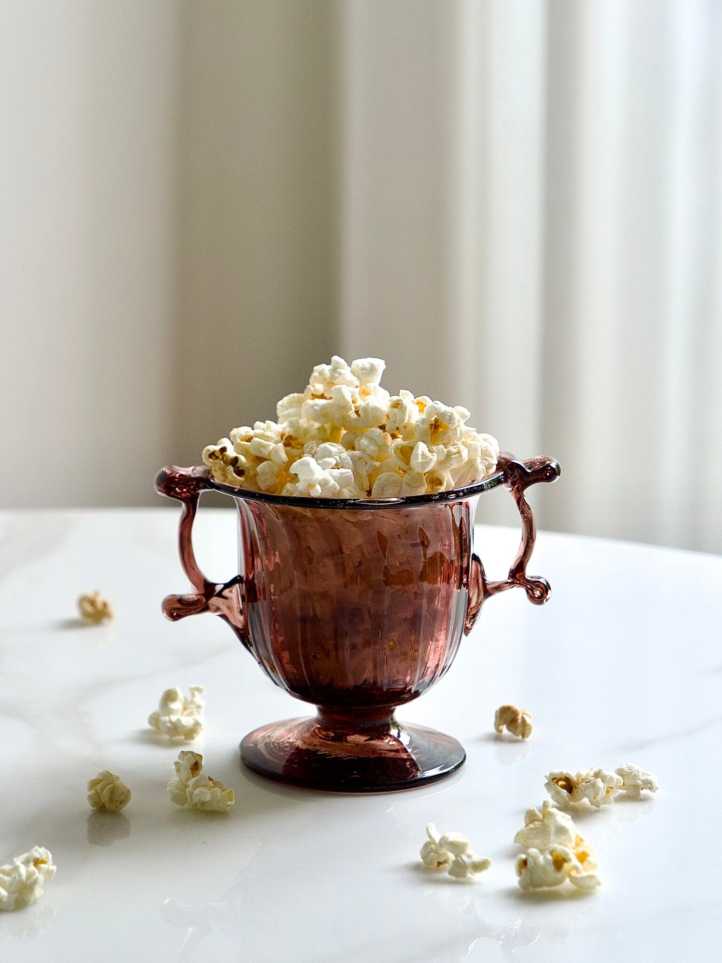 Elegant pink glass bowl for everyday use filled with popcorn on a white surface with scattered popcorn.