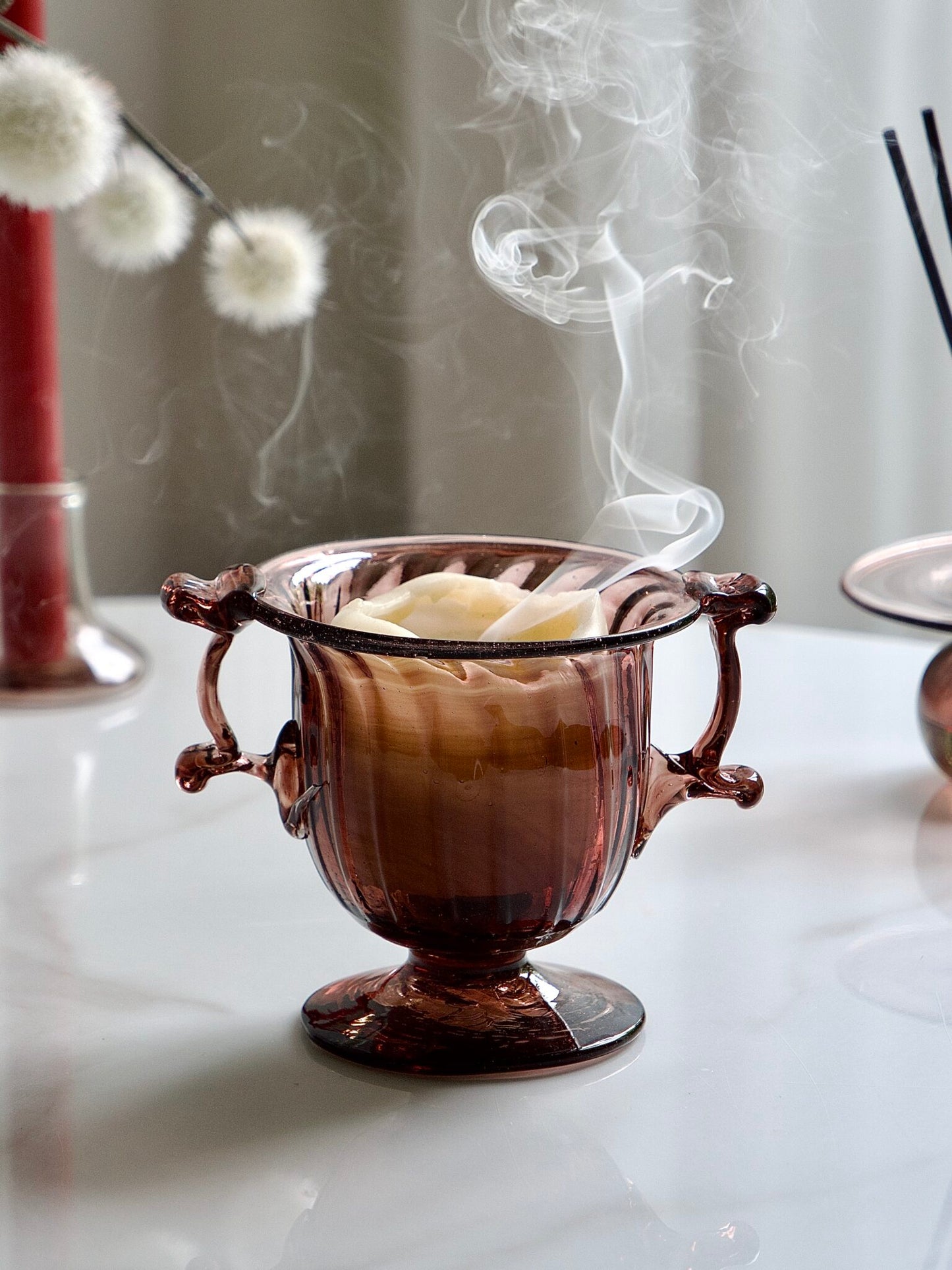 Decorative glass bowl with handles on a white surface, surrounded by flowers and candles.