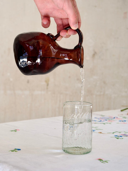 Handmade brown and raspberry-hued glass pitcher pouring water into a glass on a light-colored surface.