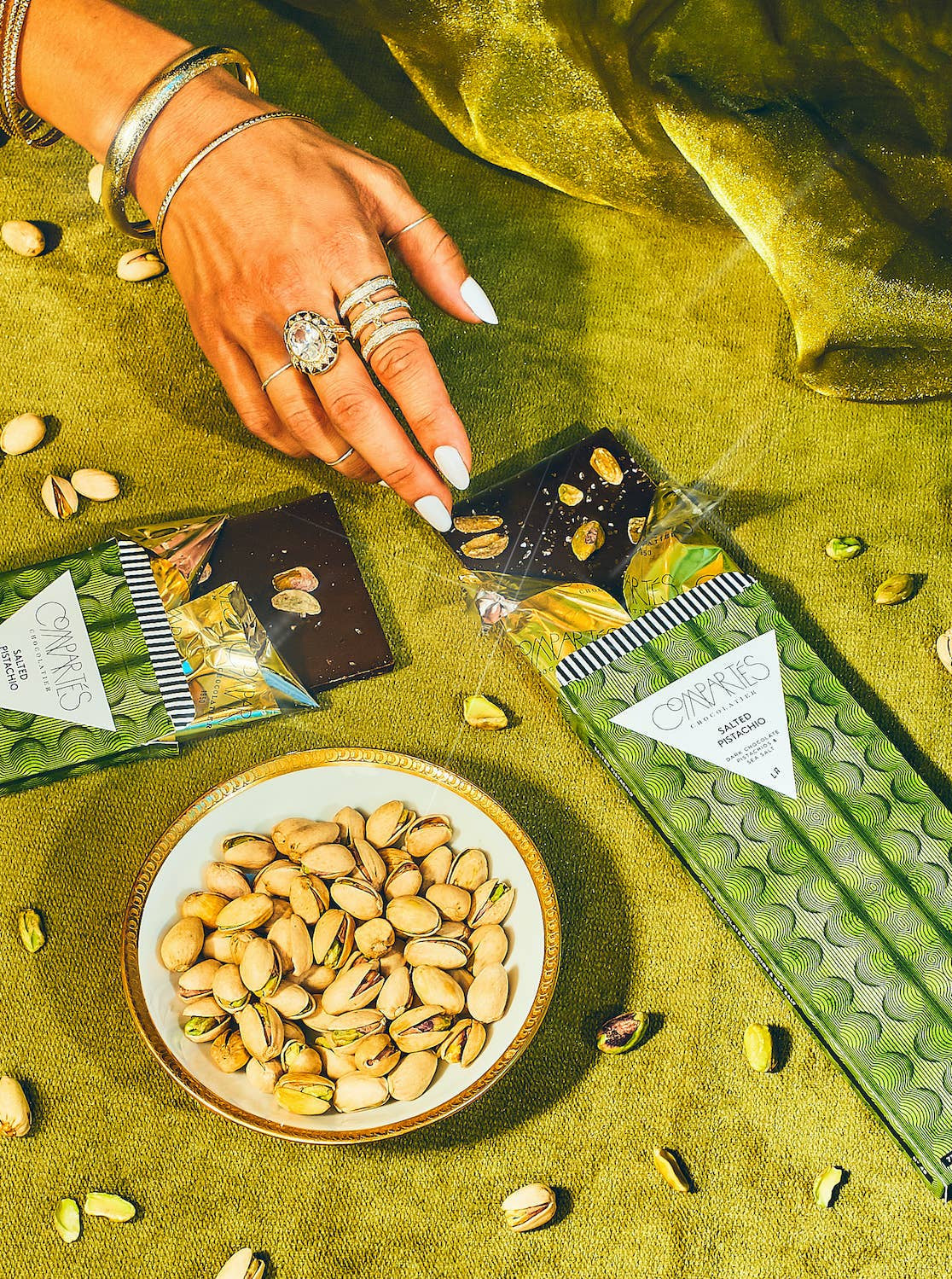 Hand reaching for a chocolate bar with pistachios on a green surface with pistachios and chocolate bars.
